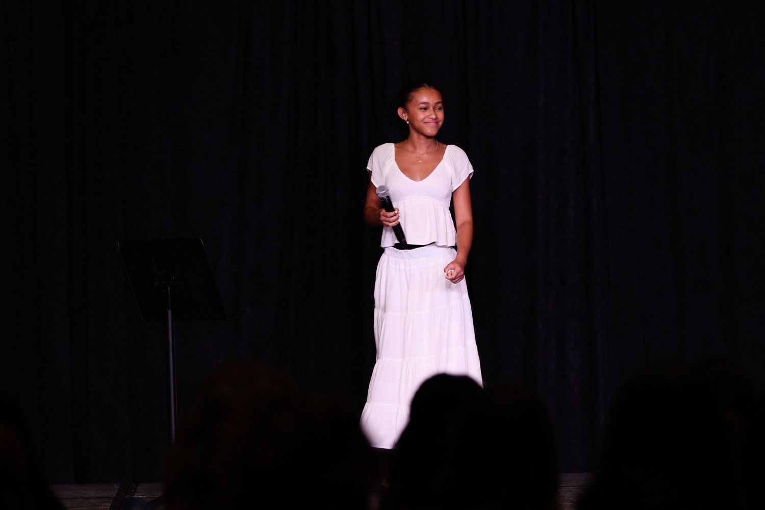 Young woman in white dress performing on stage with microphone against dark curtain backdrop.