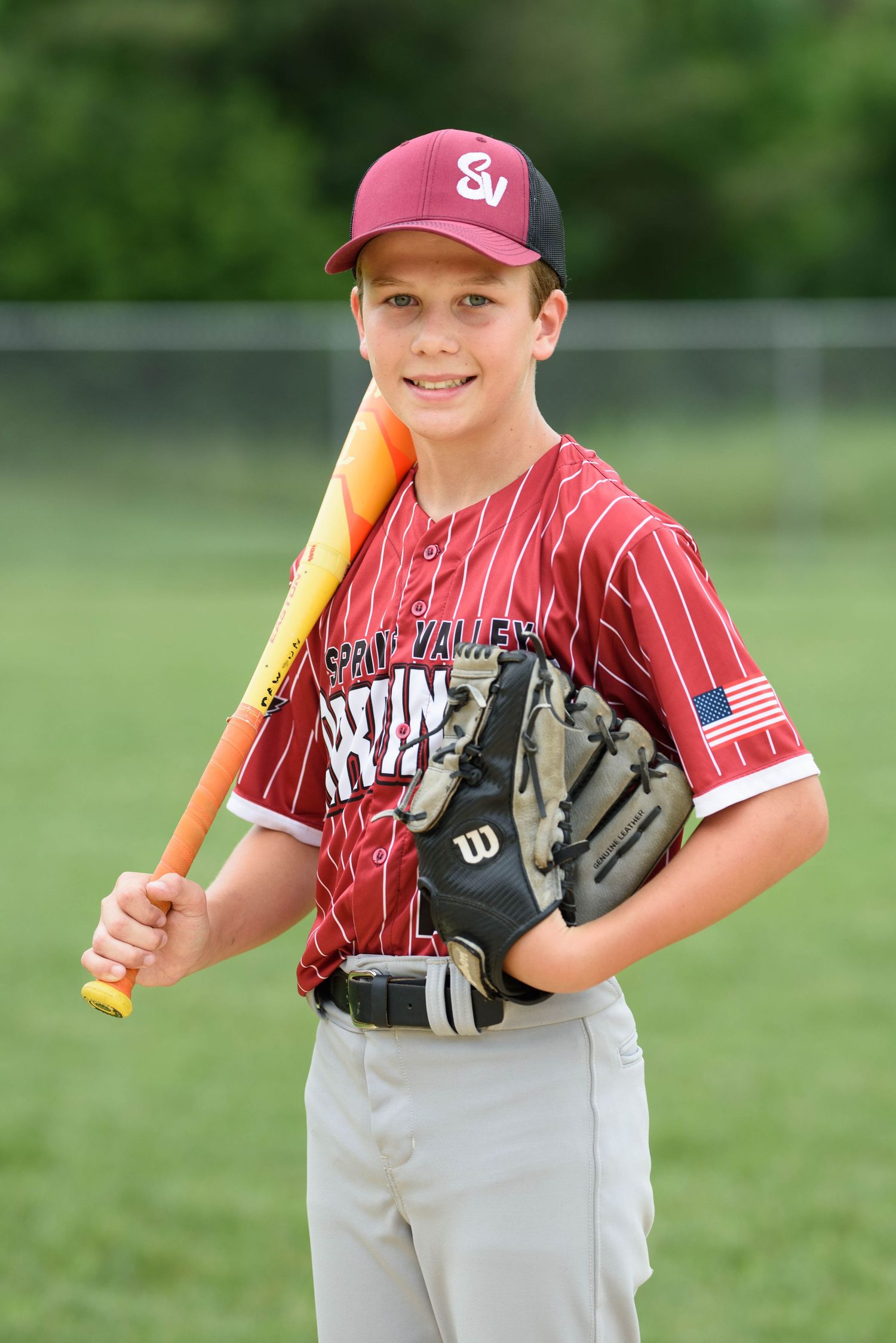 Young baseball player in maroon pinstripe uniform holding bat and Wilson glove, smiling outdoors on field.