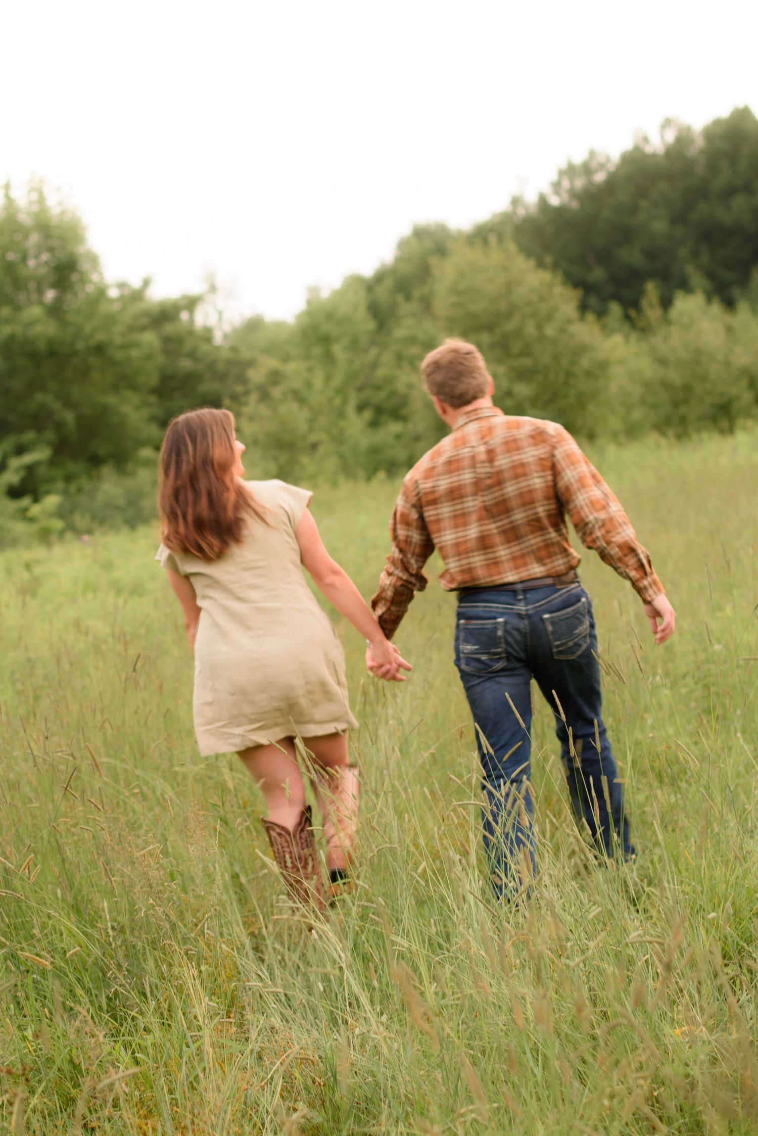 A couple holds hands while walking through a lush green meadow, the woman in a dress and boots, the man in plaid shirt and jeans.