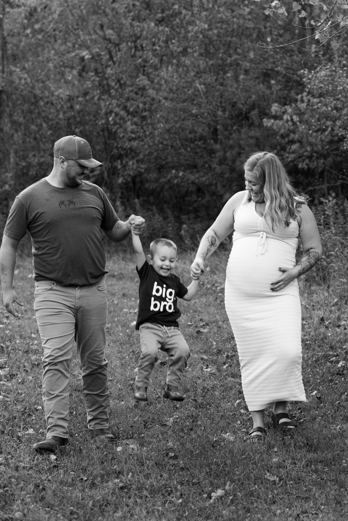 Black and white photo of pregnant woman, man, and toddler in “big bro” shirt walking outdoors.