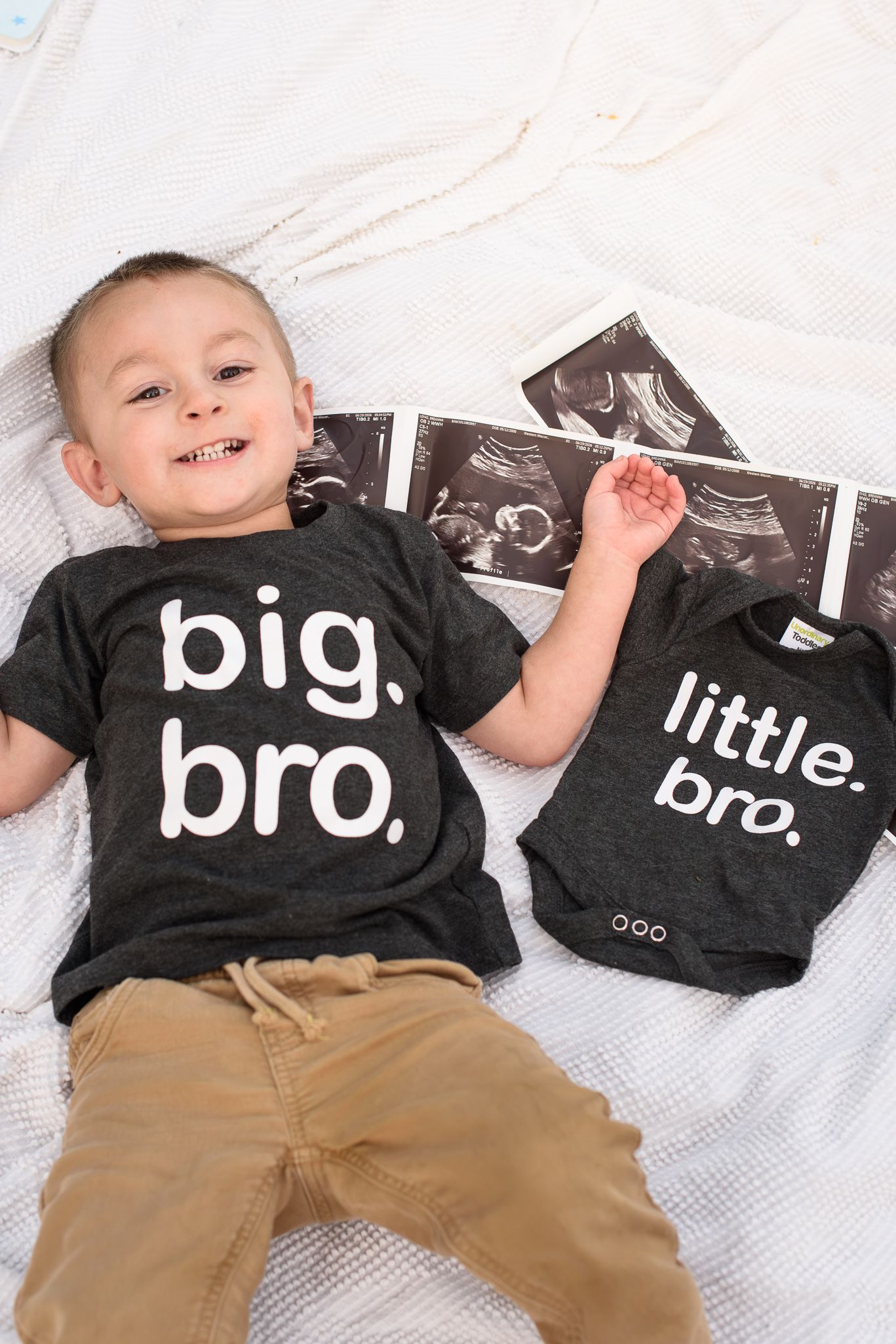 Toddler boy in big bro shirt lying next to little bro onesie and ultrasound photos announcing pregnancy.