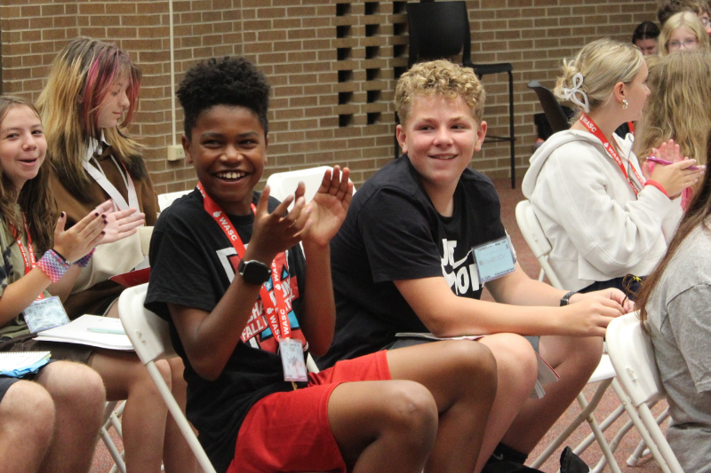 Two smiling boys clap and laugh while seated at a school or camp event indoors.