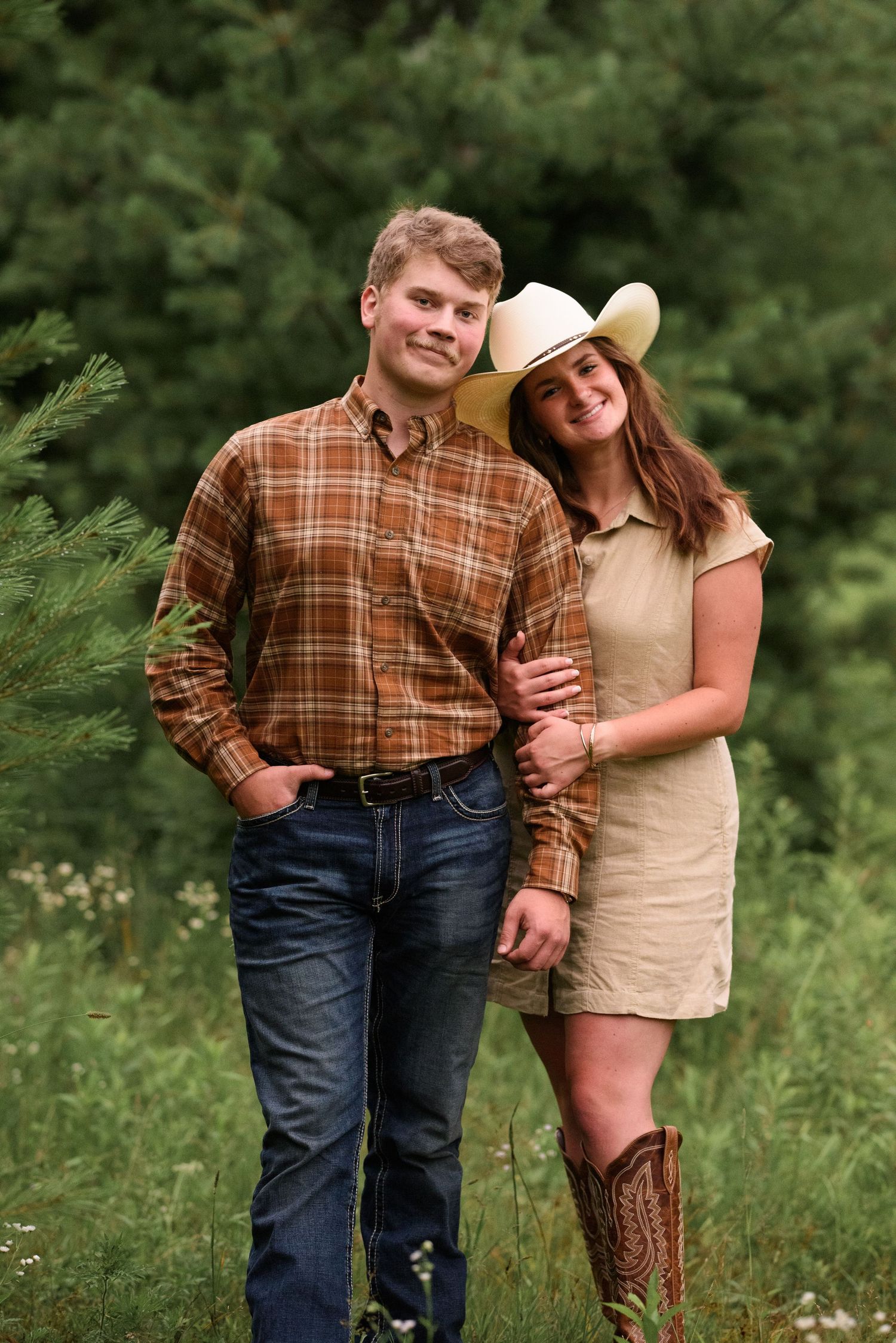 A young couple posing outdoors, woman in cowboy hat and boots leaning on man in plaid shirt.