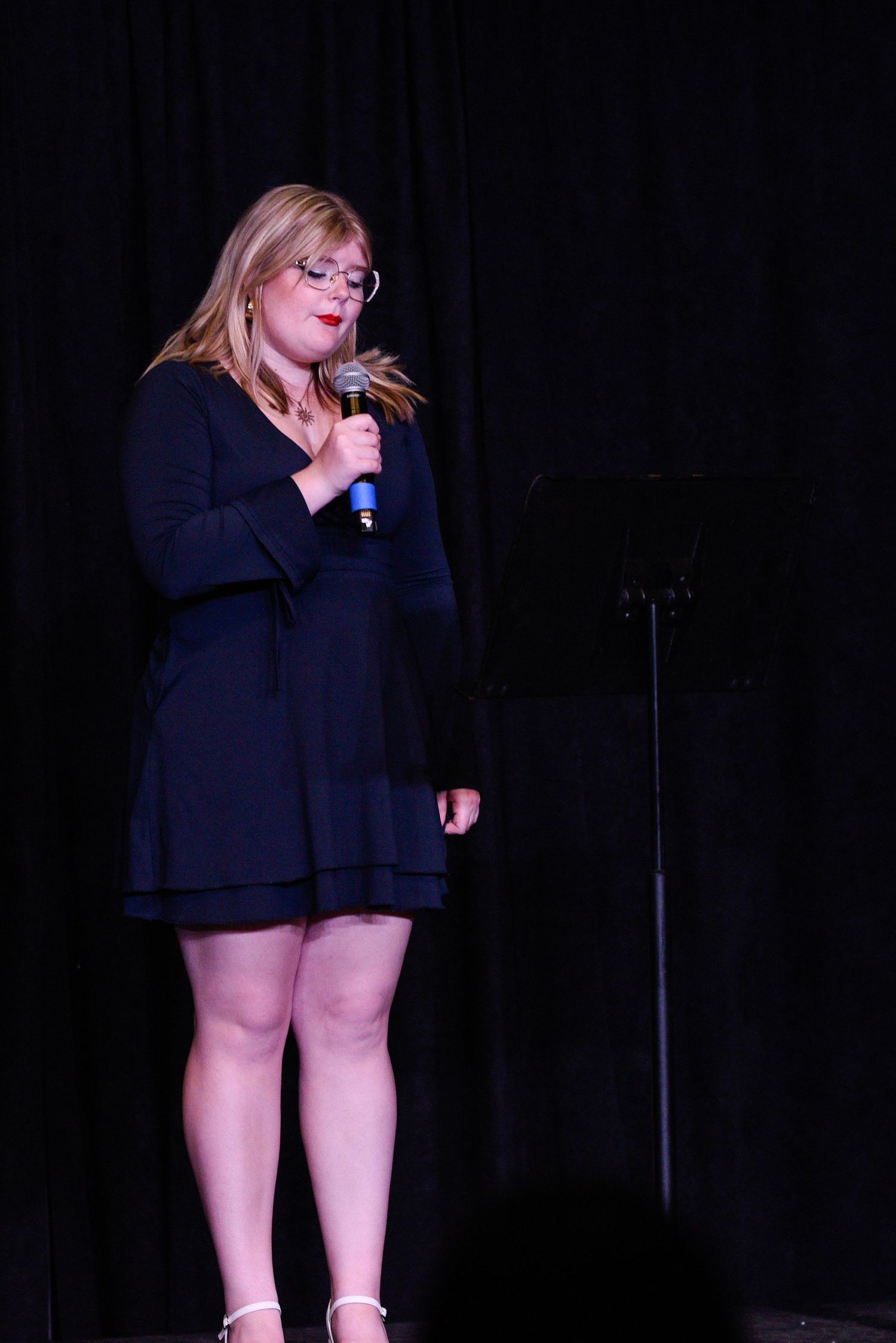 Woman in black dress singing into microphone on stage under spotlight at live performance event.