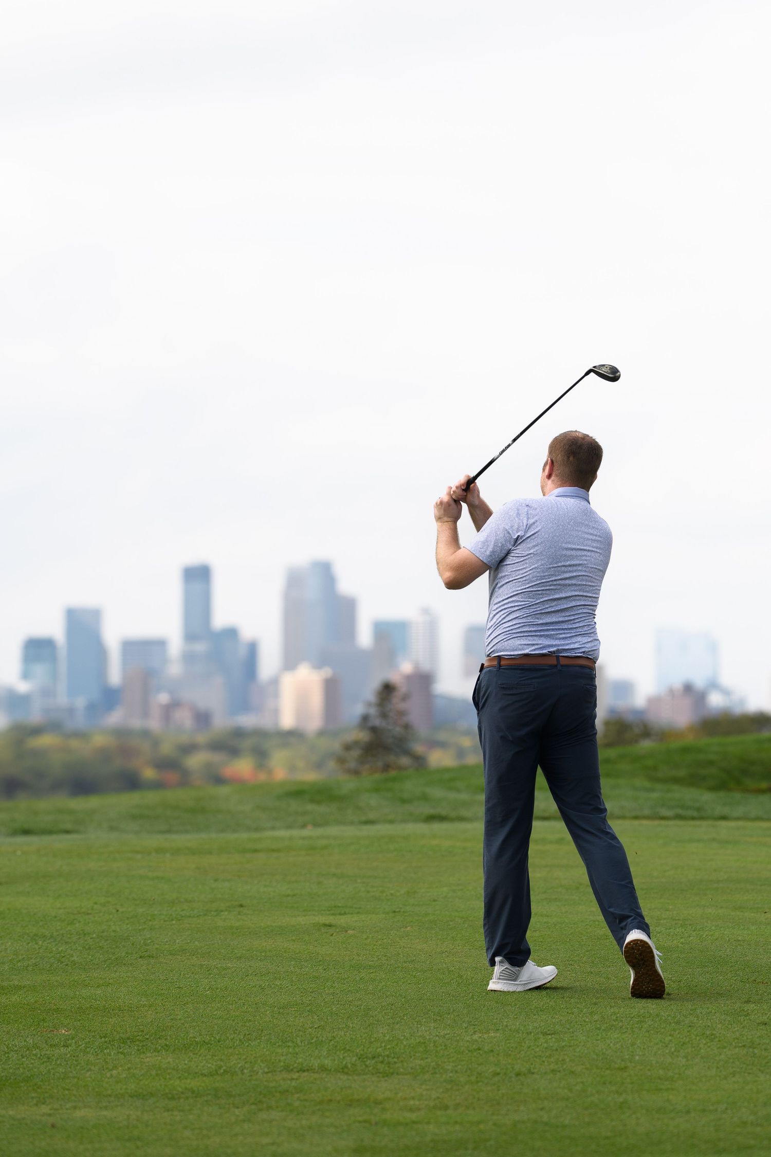 Golfer completing a swing on a lush fairway with a city skyline in the background.