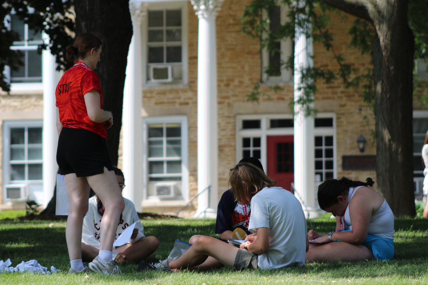 Students relax and study together on a grassy lawn outside a historic brick college building on a sunny day.