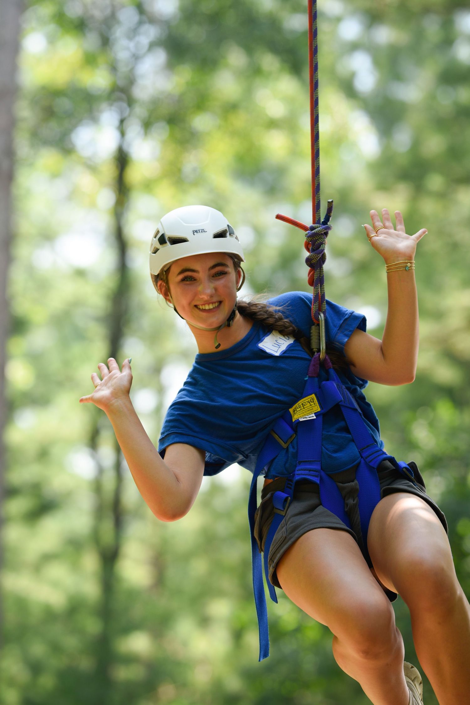 Smiling girl in blue shirt and white helmet enjoys zip-lining through green forest at summer camp.