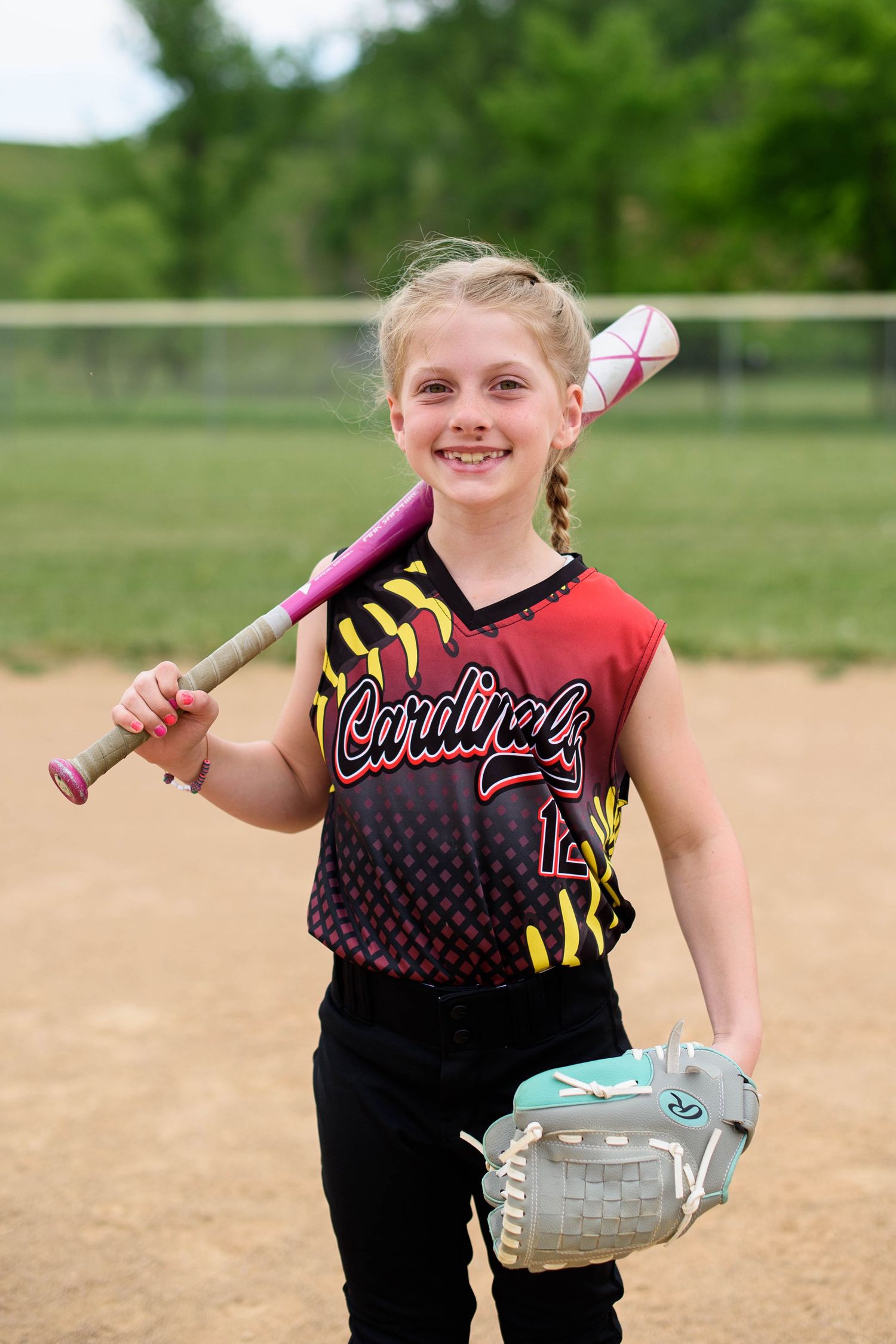 Young girl in Cardinals softball uniform smiling, holding pink bat and glove on baseball field.