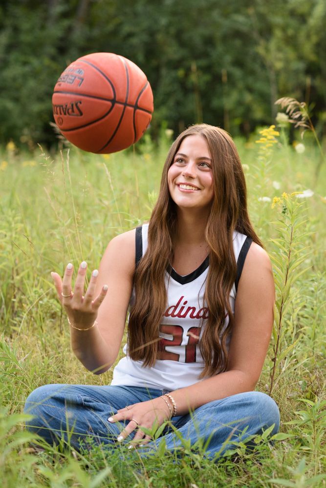 Smiling teen girl in Cardinals jersey tosses Spalding basketball while sitting cross-legged in a lush green meadow.