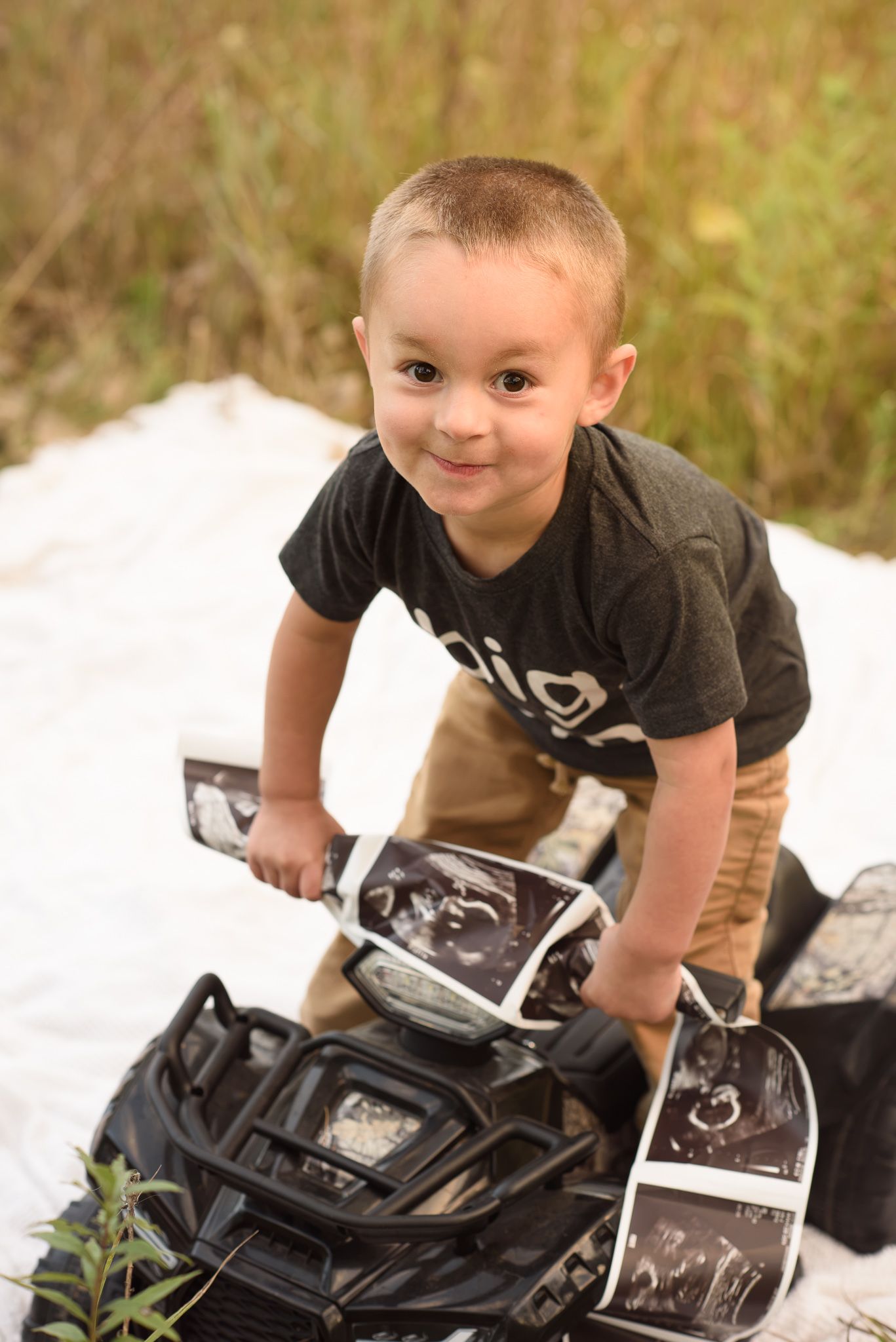 Young boy smiles while holding ultrasound photos on a toy ATV during an outdoor maternity photo shoot.