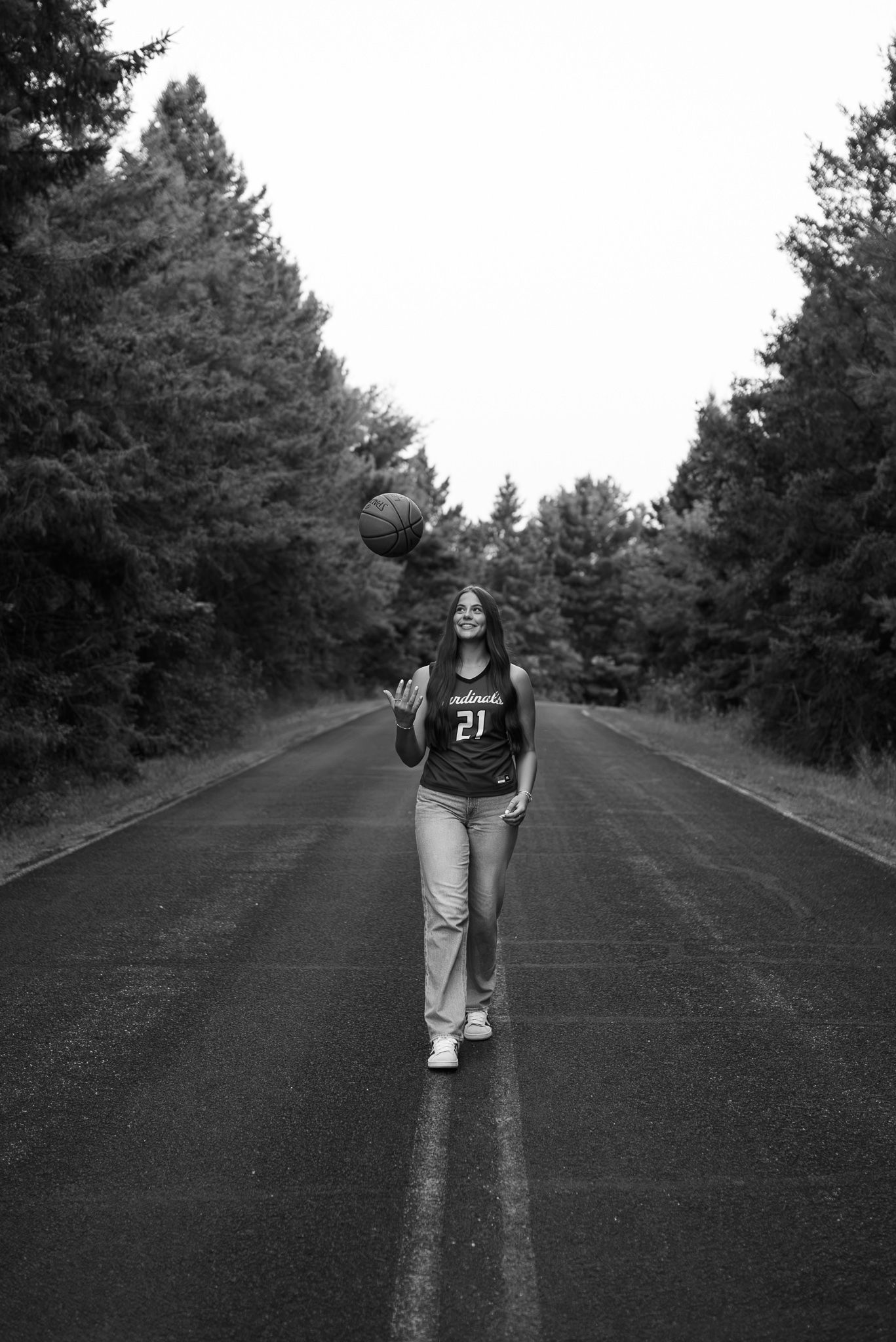 Black and white photo of a girl in jersey number 21 tossing a basketball on a tree-lined road.