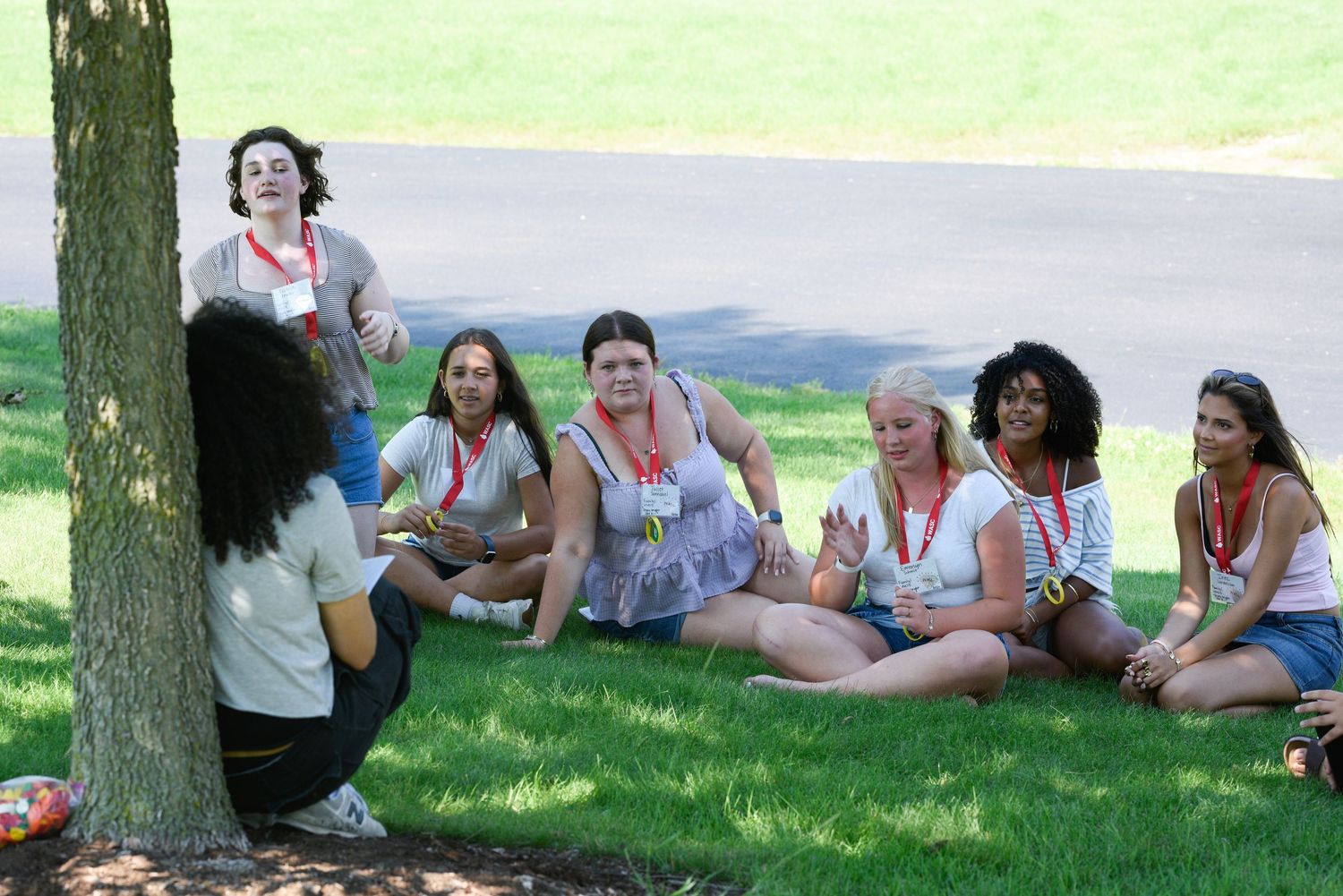 Group of women sitting on grass near a tree outdoors, enjoying a sunny day at a park or campus.