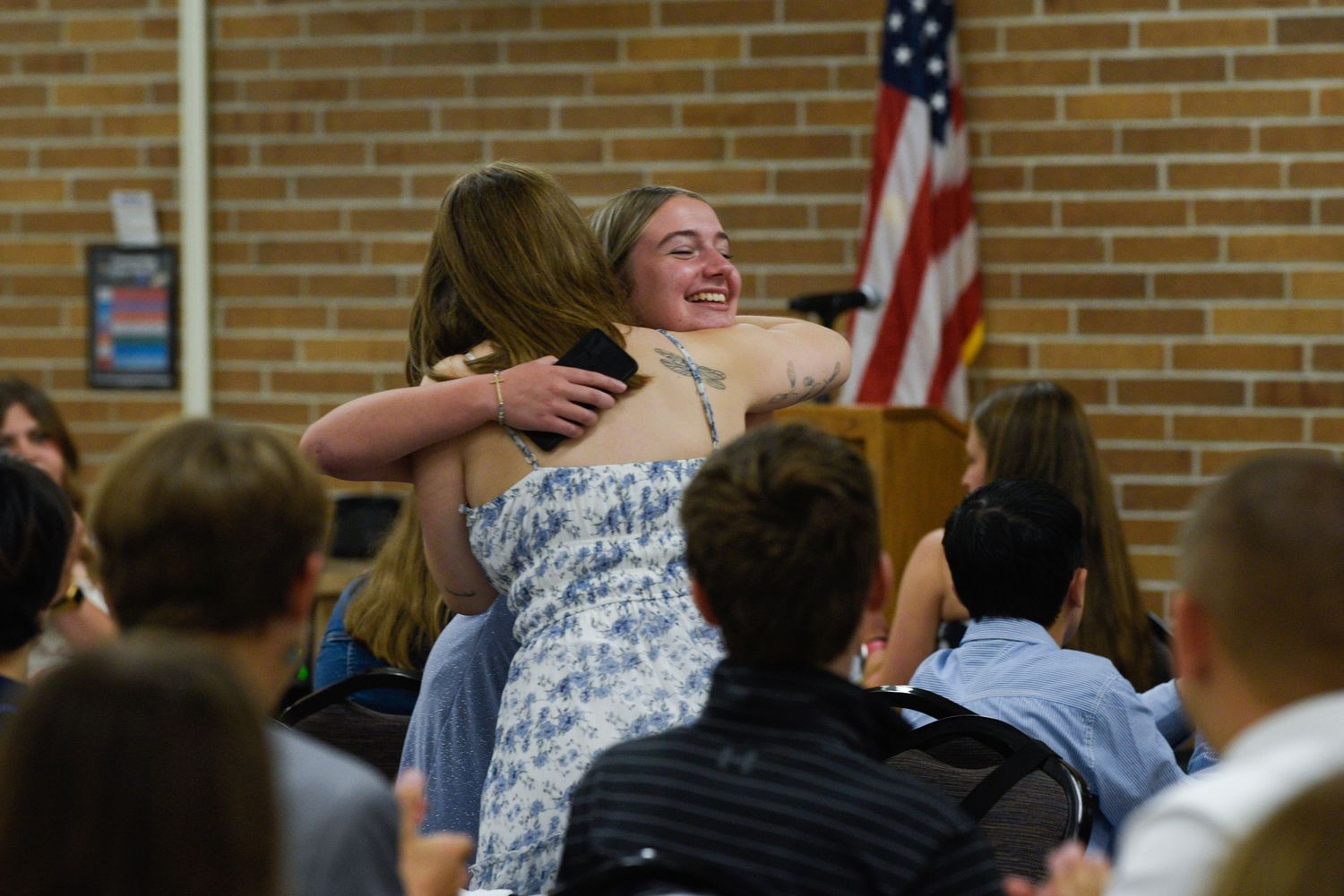 Students hugging and celebrating in a school auditorium with an American flag in the background.
