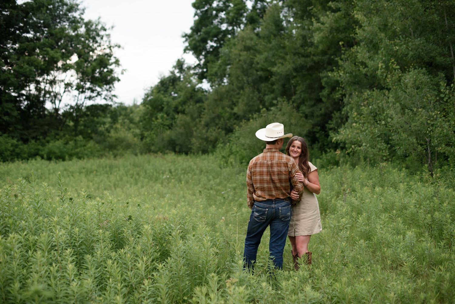 Couple embracing in a lush green field, man wearing cowboy hat and plaid shirt with woman in beige dress.