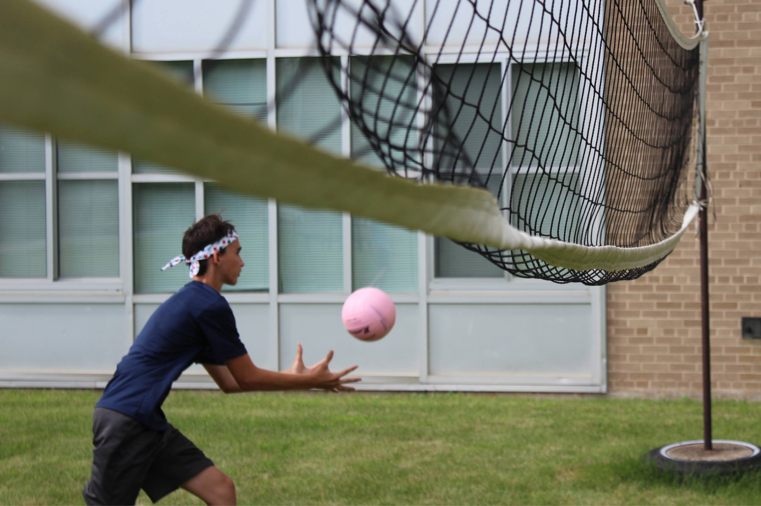 A student wearing a headband bumps a pink volleyball over a net during an outdoor game.