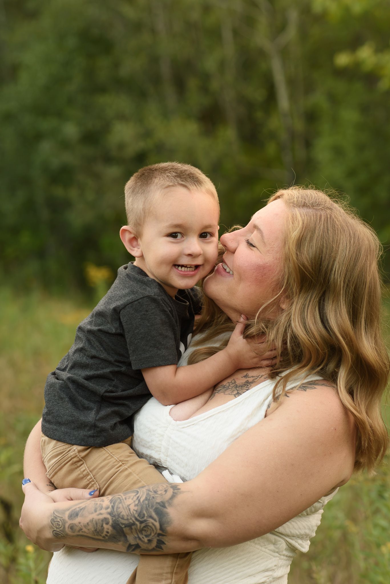 A woman with tattoos holds and receives a kiss from a smiling toddler boy outdoors in a green nature setting.