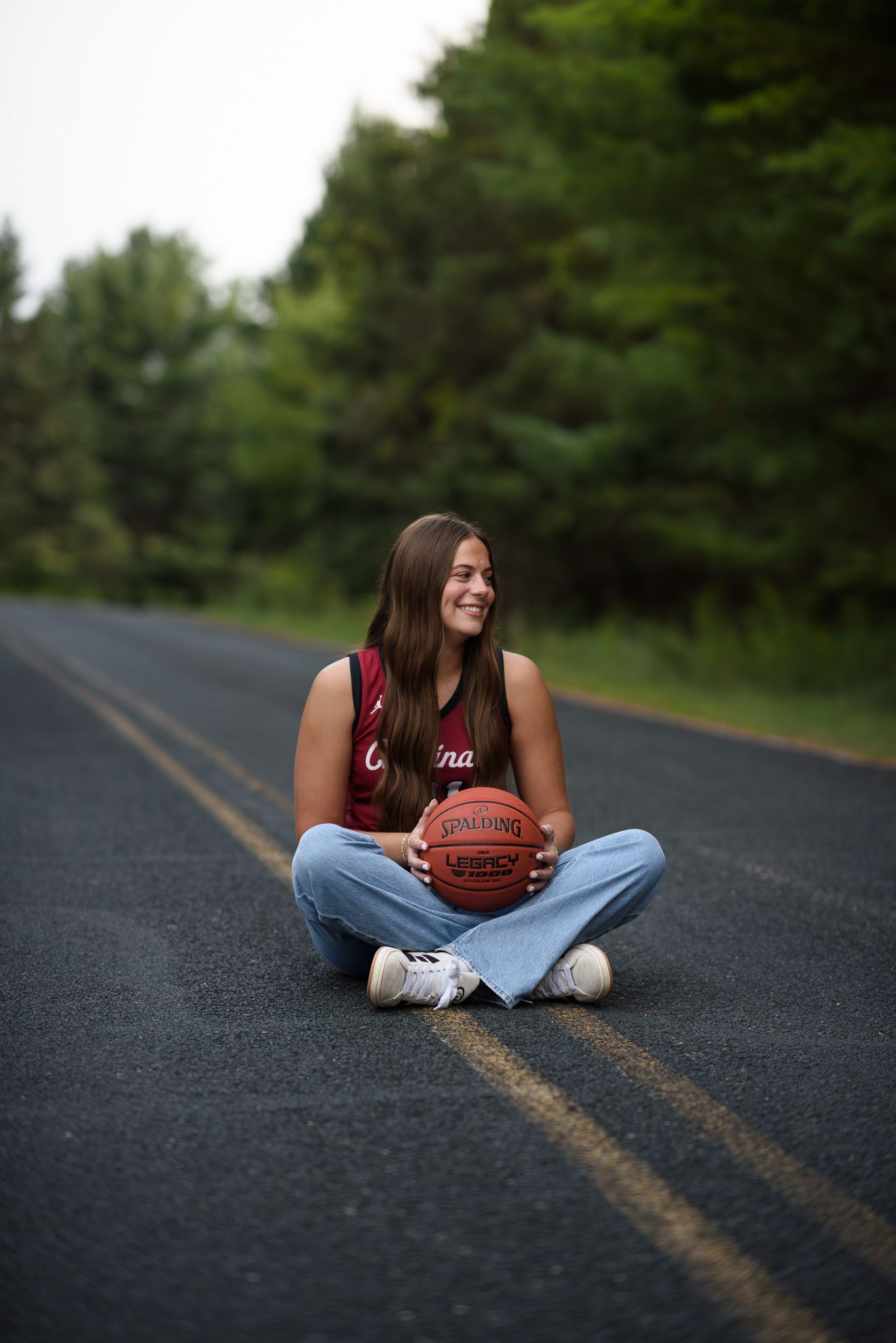 Smiling girl in Cardinals basketball jersey sitting cross-legged on a road holding a Spalding basketball.
