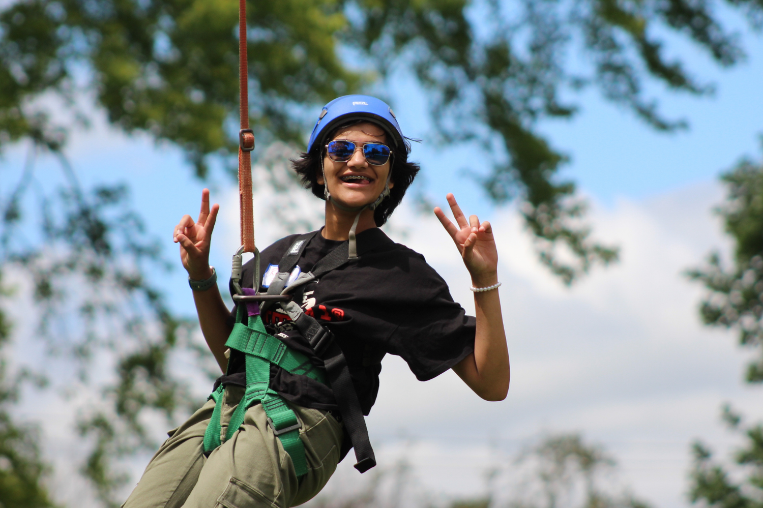 A person wearing a blue helmet and harness makes peace signs while zip-lining through green trees.