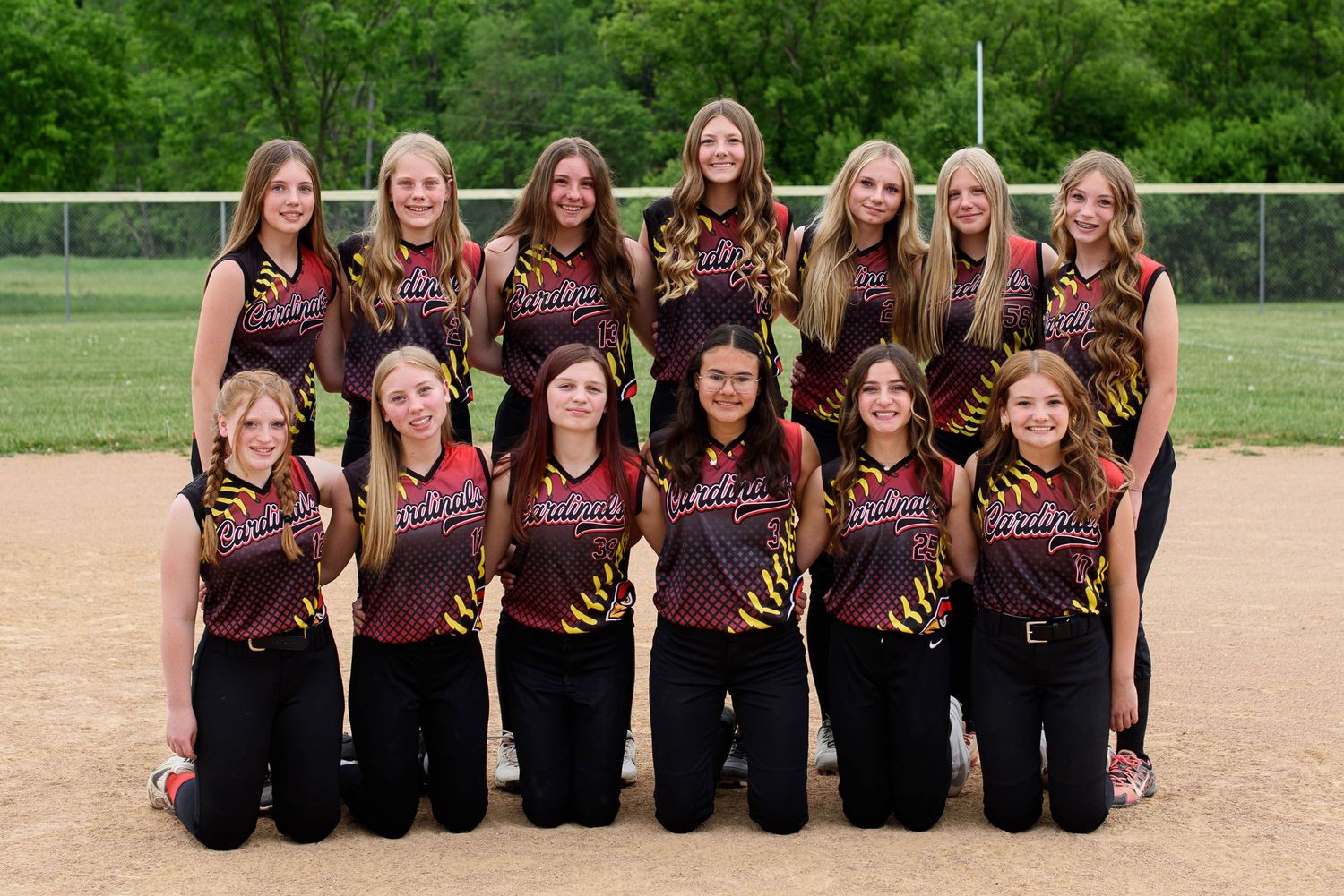 Girls softball team posing for group photo in matching red and black tiger-striped uniforms on a dirt field.