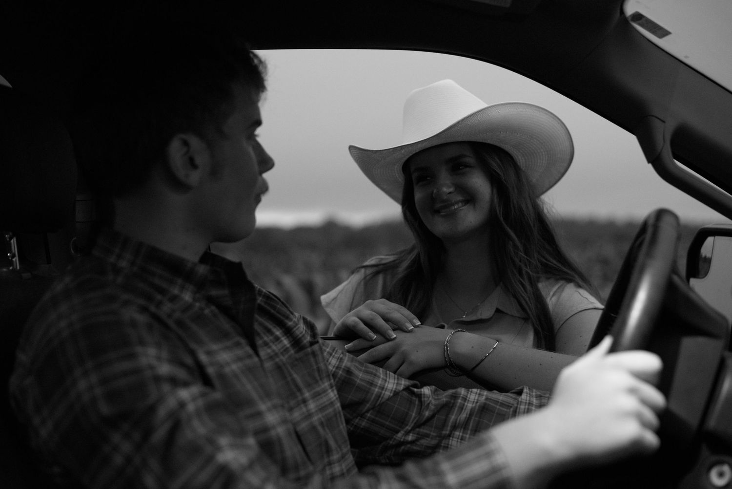 Black and white photo of a couple sitting in a car, woman wearing a cowboy hat smiling at a man in a plaid shirt.