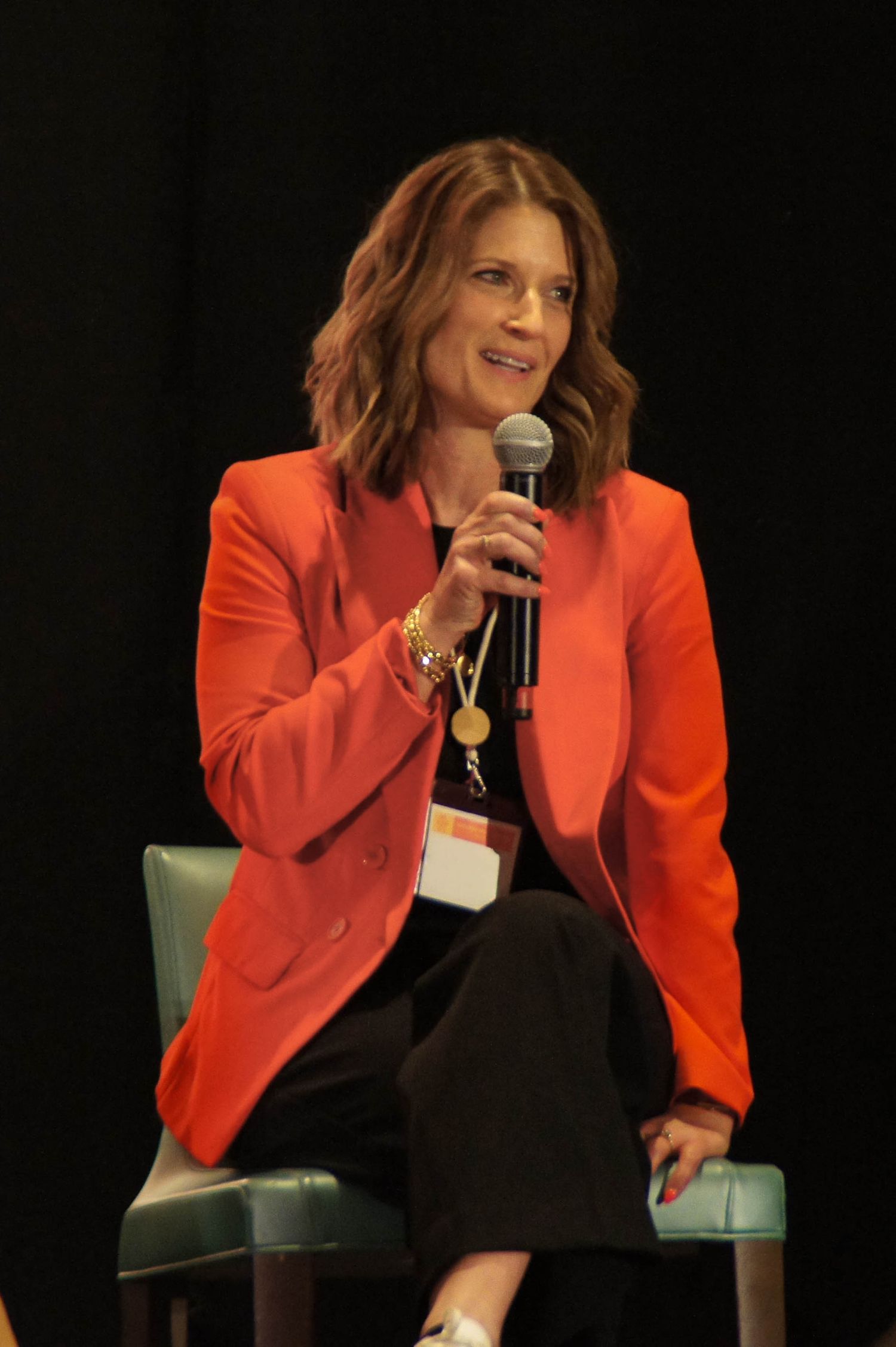 Woman in red blazer speaks into microphone at a professional conference panel discussion.