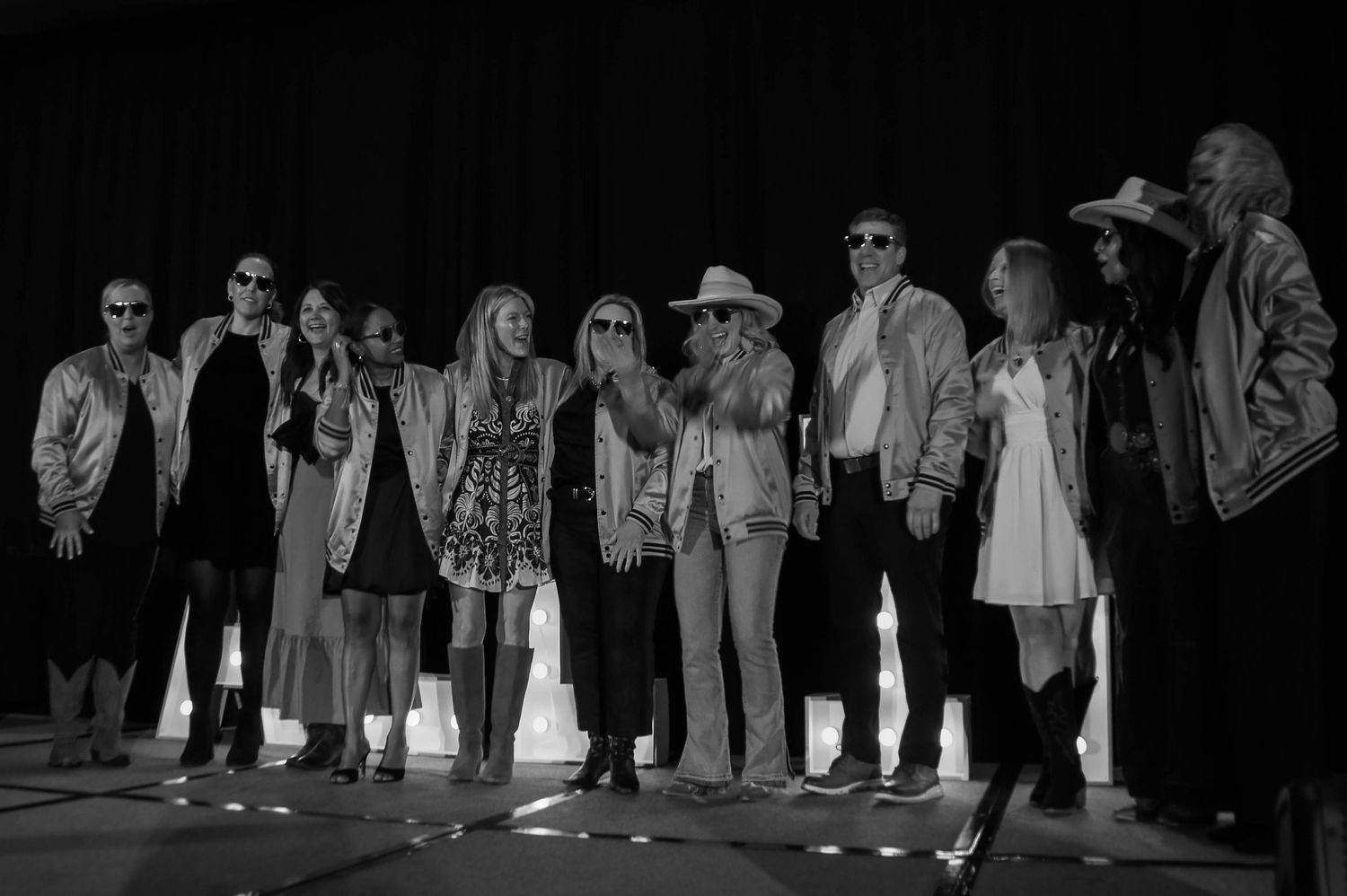 Black and white photo of large group of people posing together on stage at ALL IN conference event.