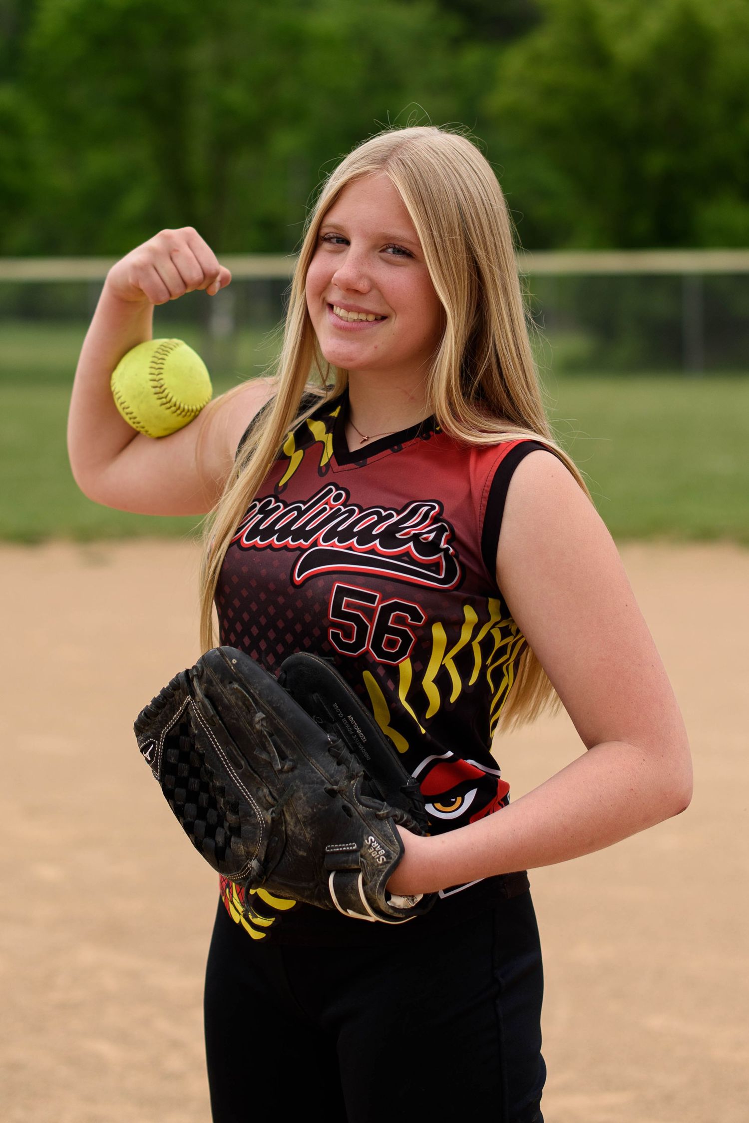 Smiling teen softball player in Cardinals jersey #56 posing with ball and glove on field.