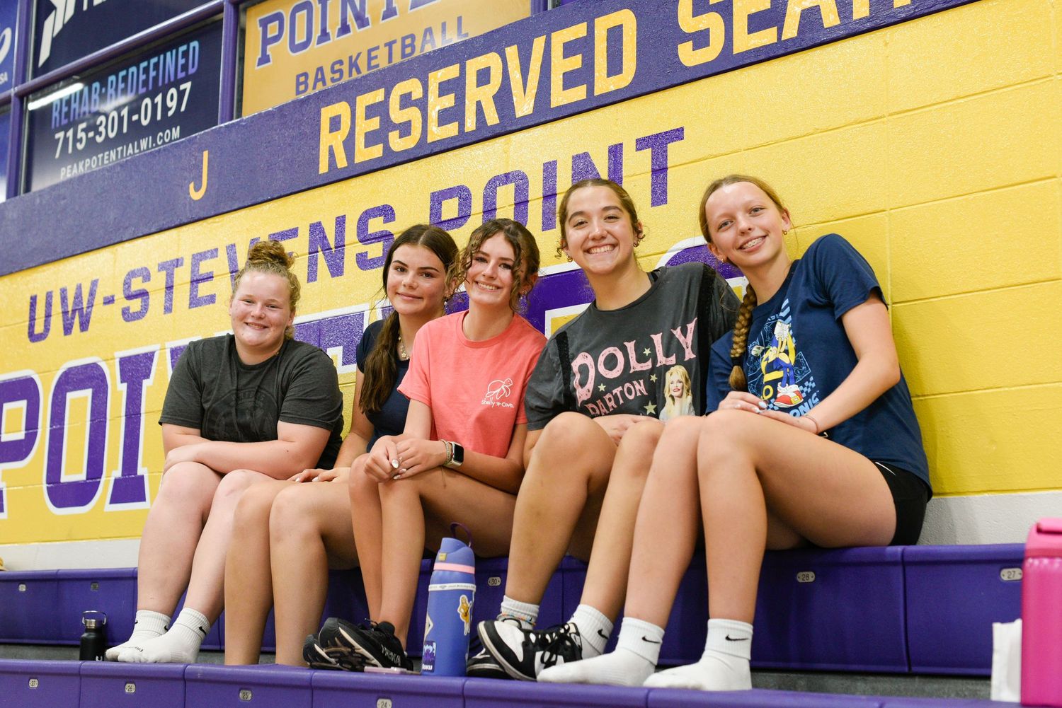 Five young women smile together in UW-Stevens Point basketball gym bleachers, wearing casual athletic wear.