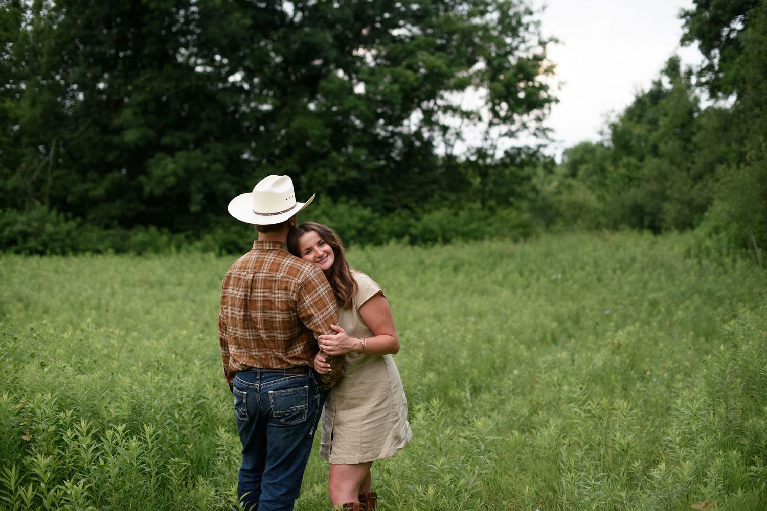 Couple embracing in a lush green field, man wearing a white cowboy hat and plaid shirt with woman in a beige dress.