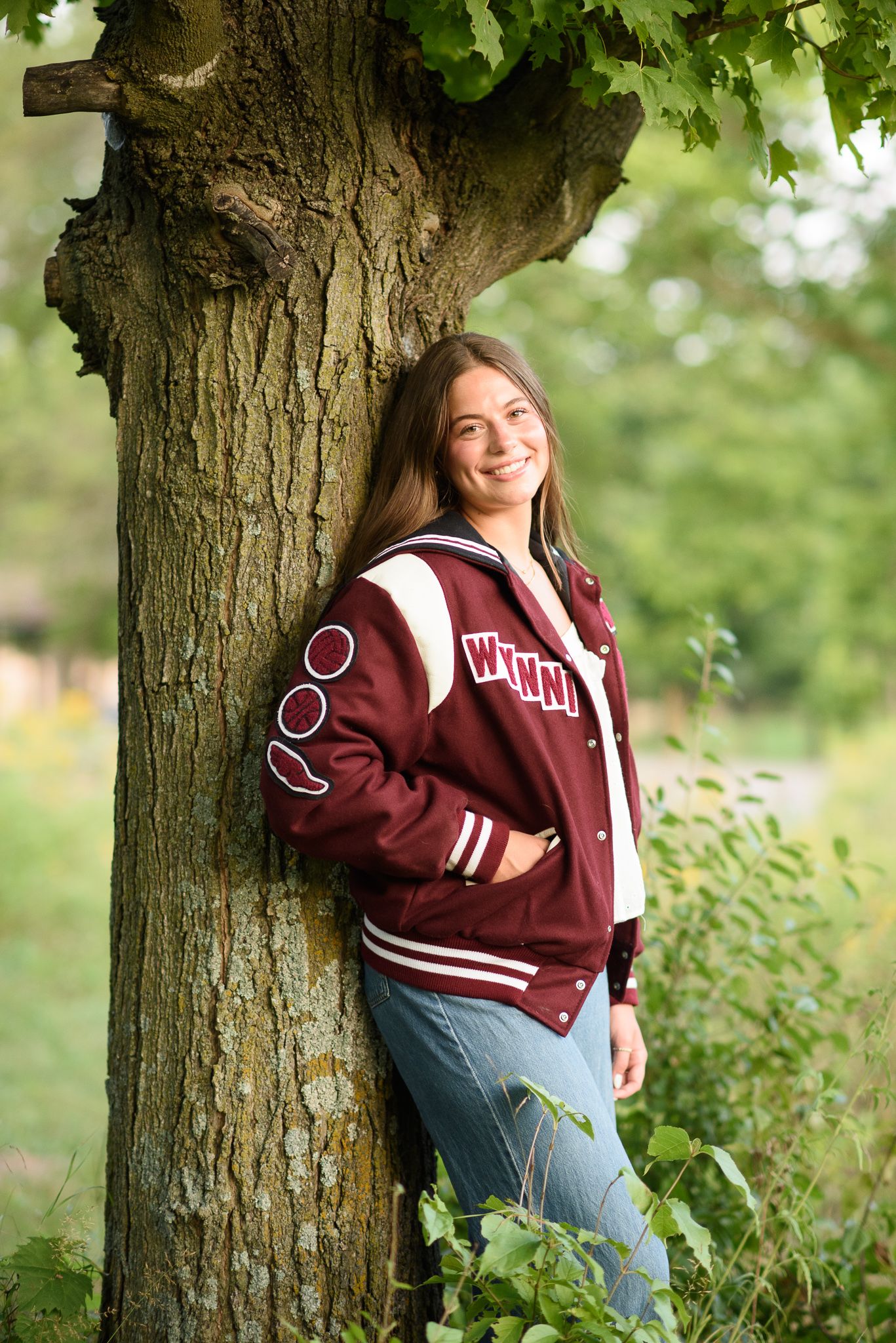 A smiling teenage girl in a maroon varsity jacket leans against a tree outdoors in a lush green setting.