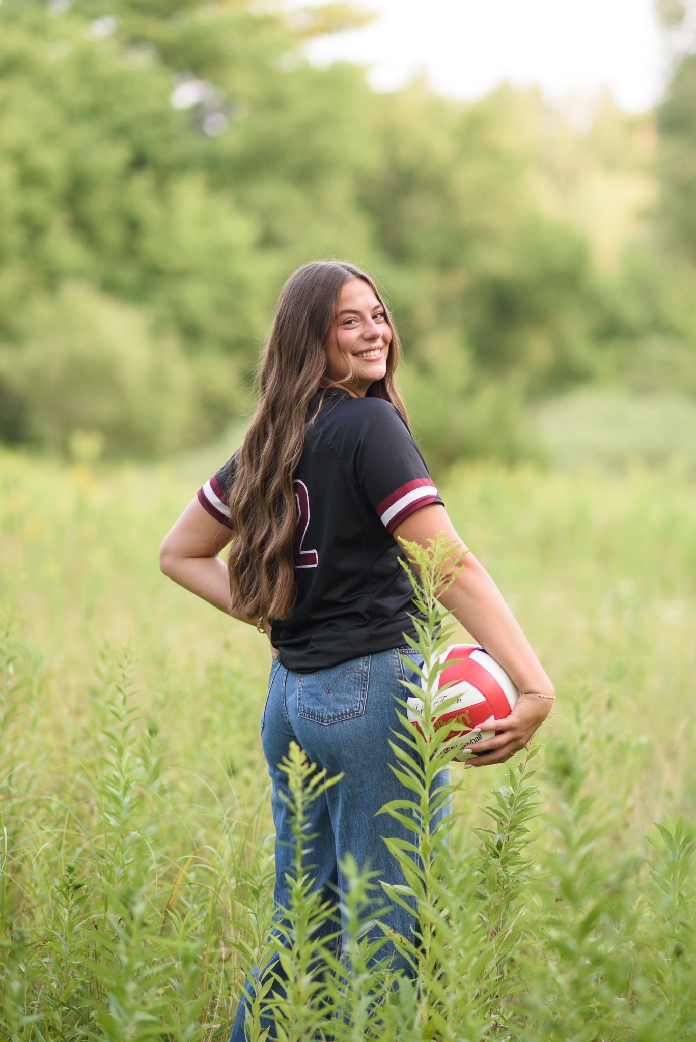 High school senior girl holding volleyball in tall grass wearing Cardinals black jersey and jeans.