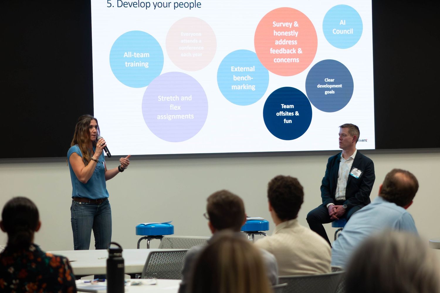 Business presenter speaks to audience beside seated colleague during corporate training seminar with slide presentation.