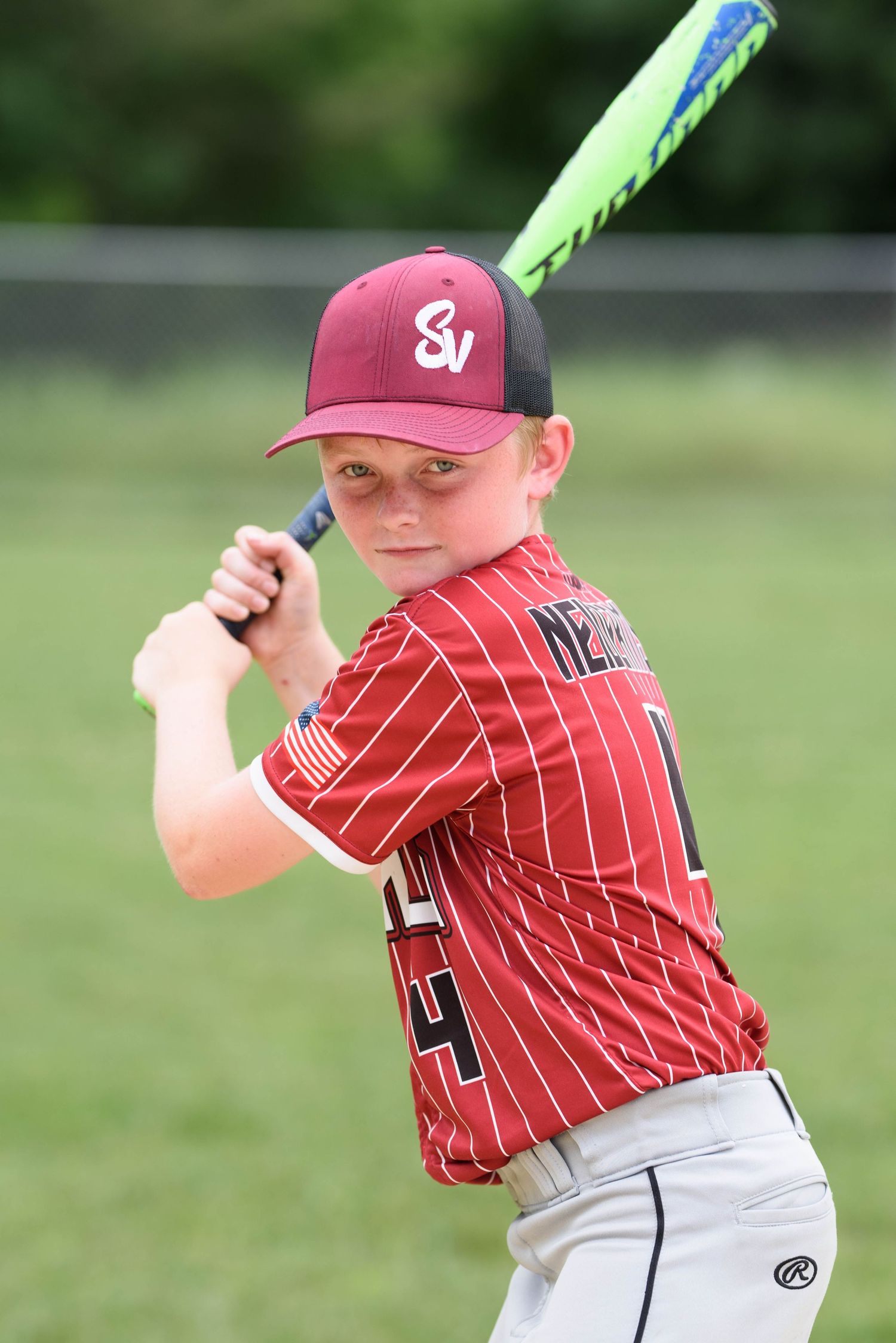 Youth baseball player in red SV Cardinals pinstripe jersey assumes batting stance with colorful bat on field.