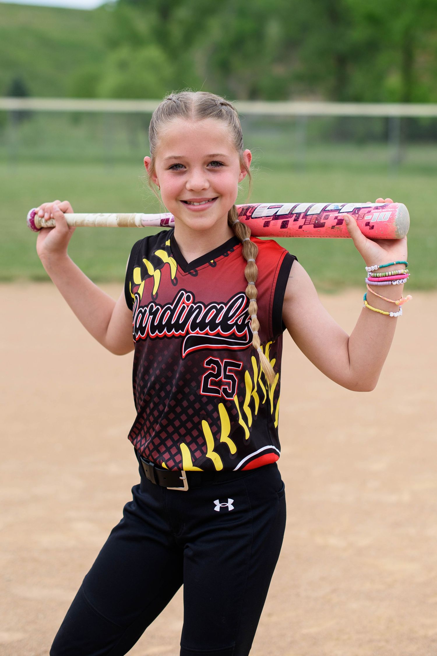 Young girl in Cardinals #25 softball uniform poses with pink bat on her shoulders on a dirt field.