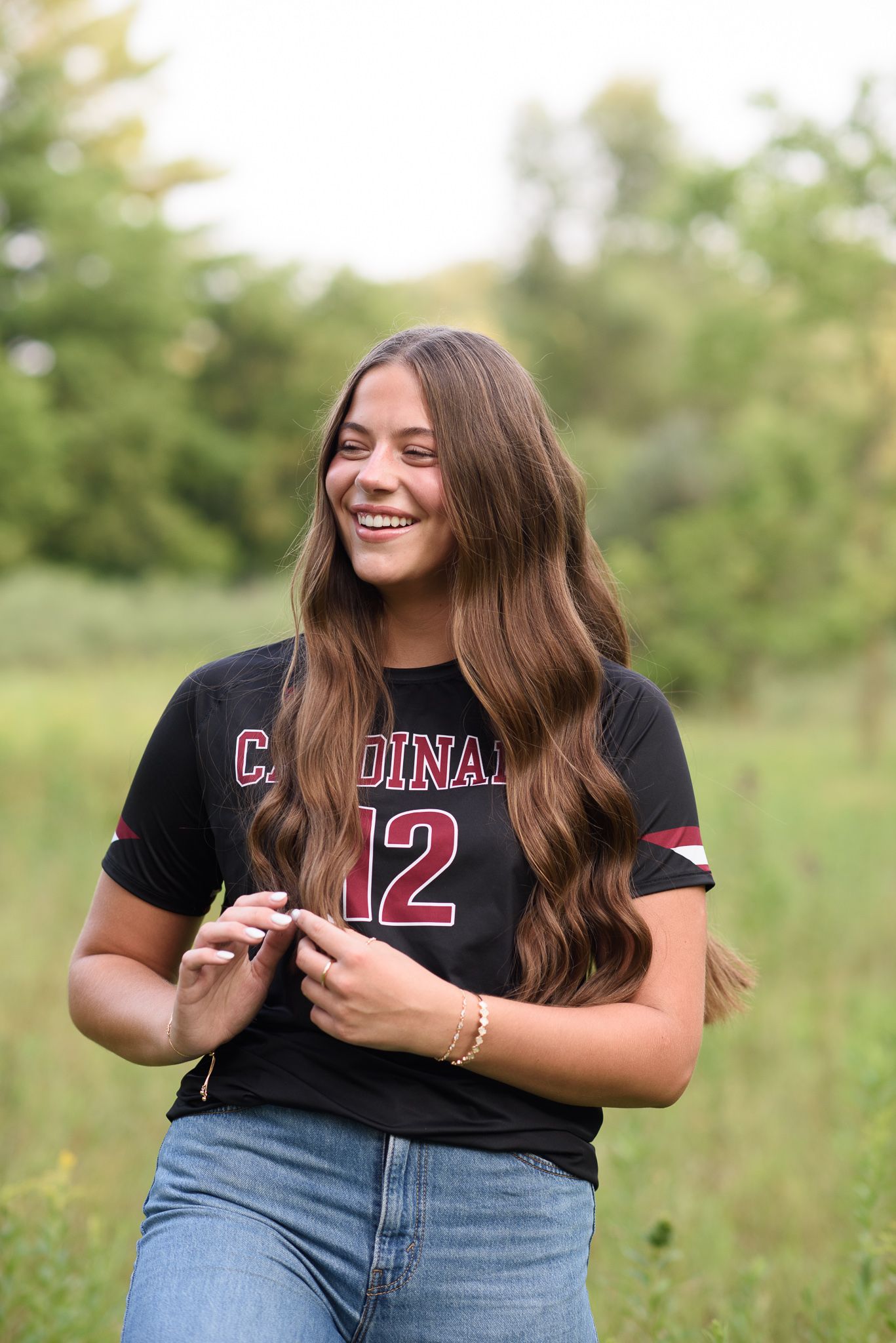 Smiling teen girl in Cardinals number 12 black jersey standing in green field during senior photo session.