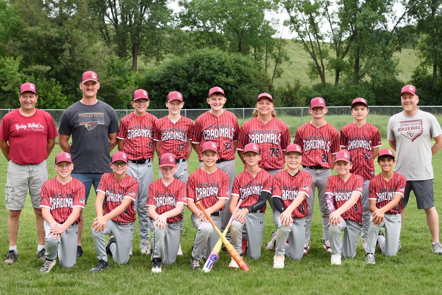 Youth baseball team in red uniforms posing for group photo outdoors with coaches and bats on green grass.