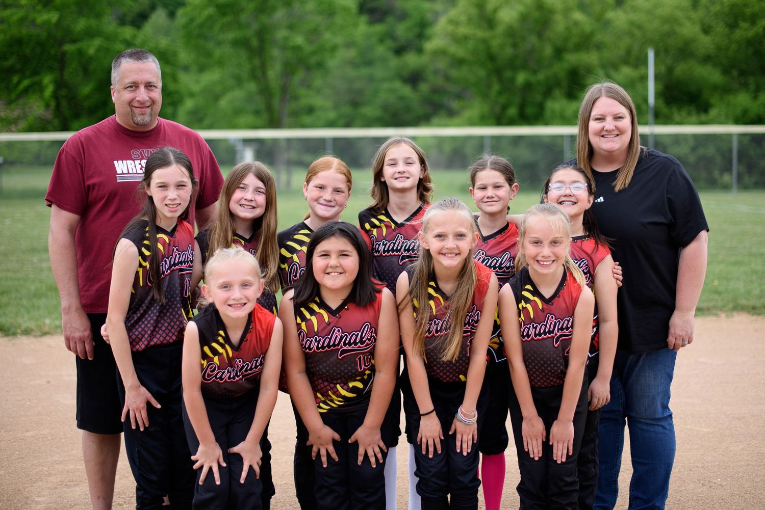 Cardinals youth softball team posing with two coaches on a baseball field in matching uniforms.