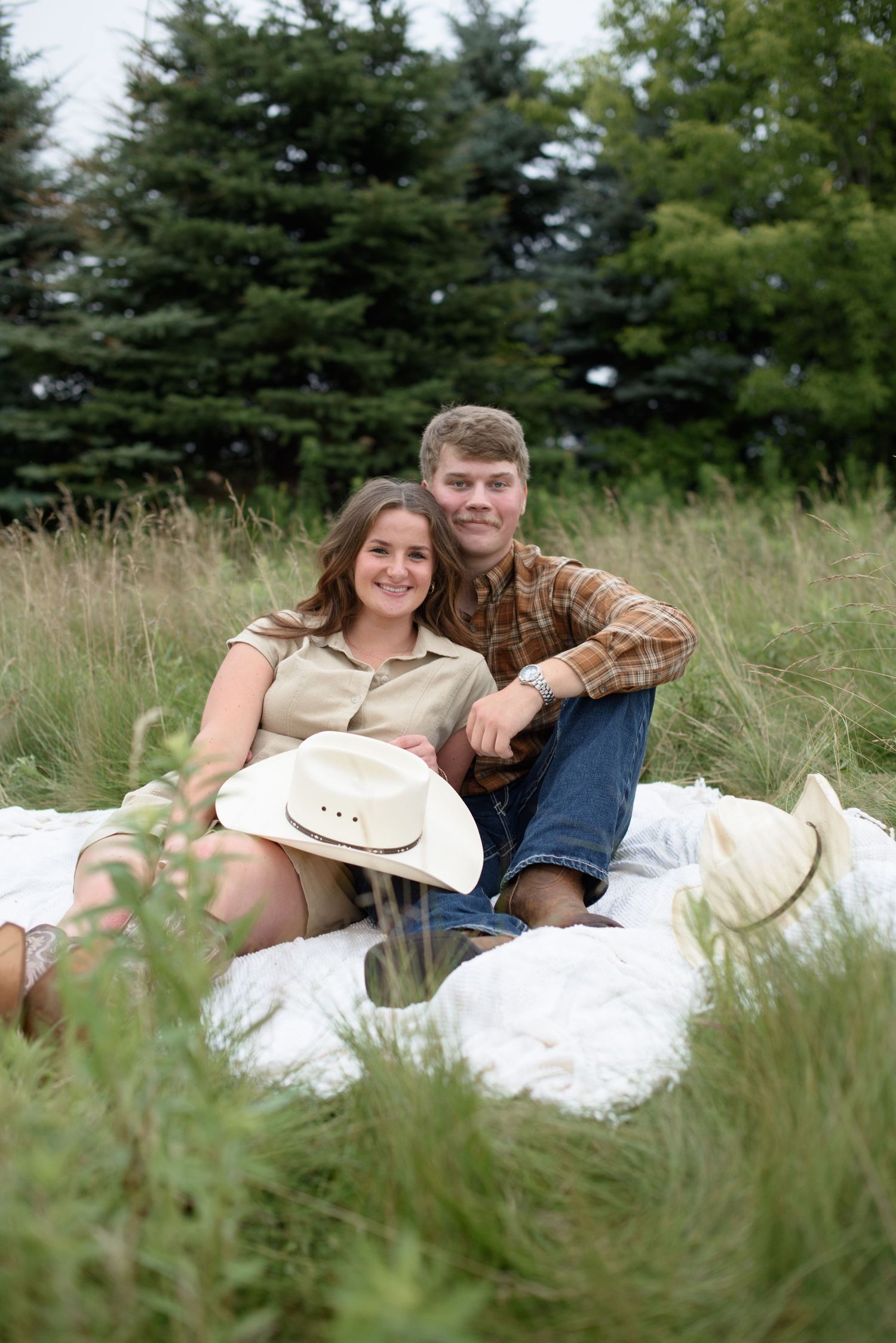 Couple sitting on white blanket in tall grass field, woman holding cowboy hat, man in plaid shirt, smiling at camera.