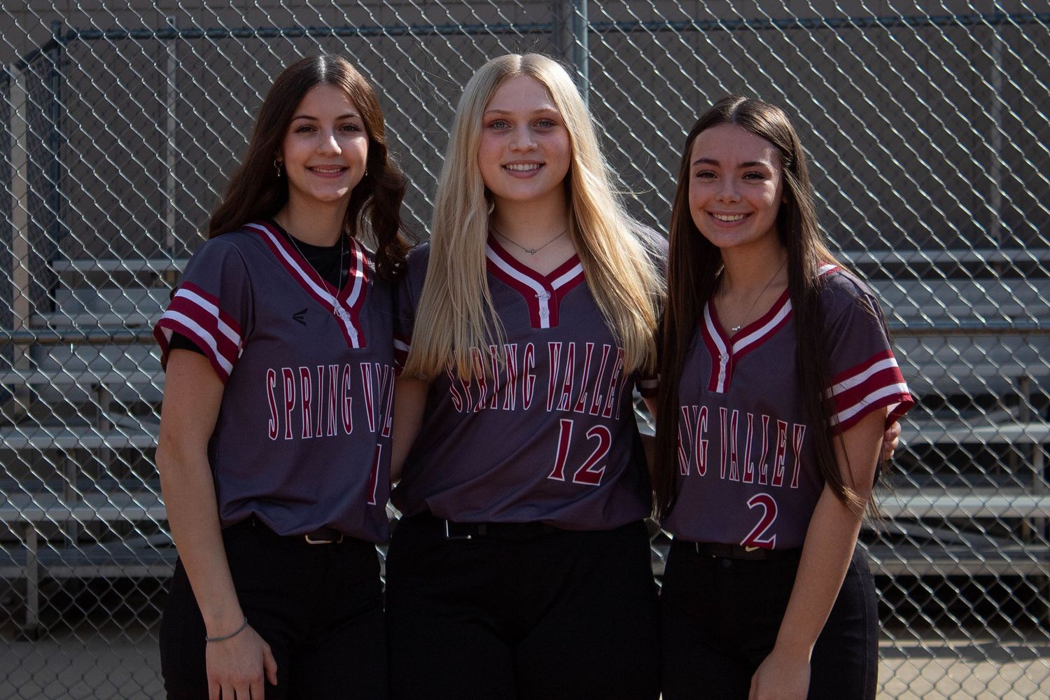 Three Spring Valley softball players pose together in maroon uniforms on dirt field.