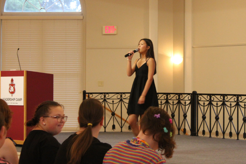 Young woman in black dress performs singing on stage before a seated audience indoors.