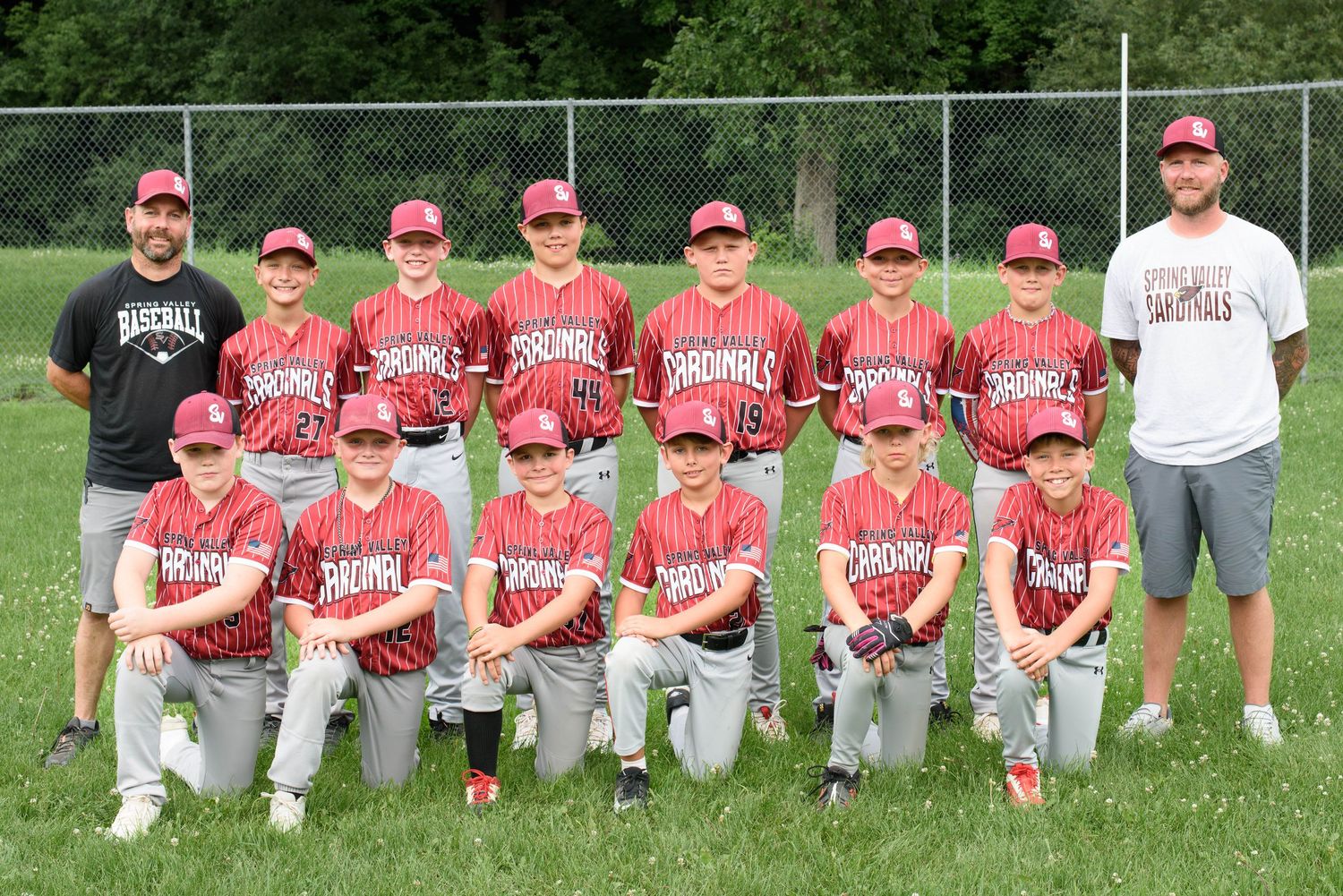 Youth baseball team in red Cardinals uniforms poses for group photo with two coaches on a green field.