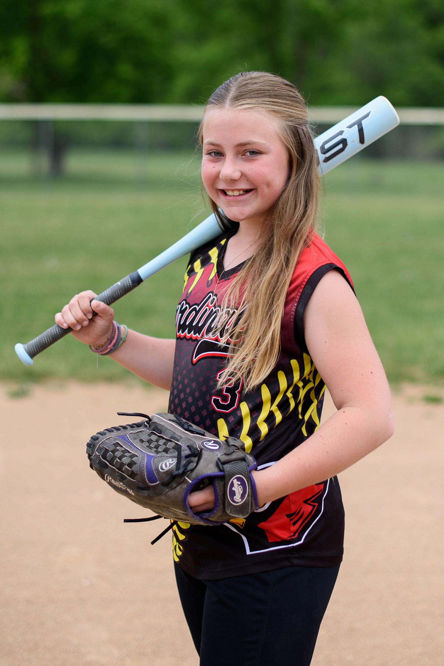 Young softball player smiling with bat over shoulder and glove in hand wearing Cardinals jersey on dirt field.