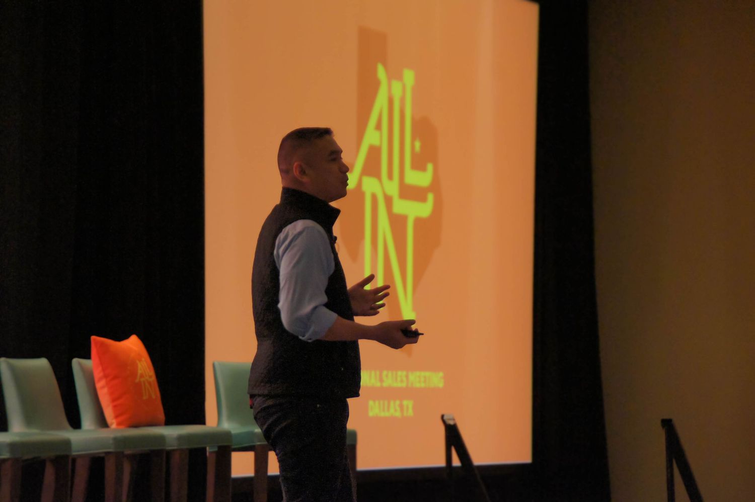 Man presenting at All In sales meeting in Dallas TX with colorful orange projection screen behind him.