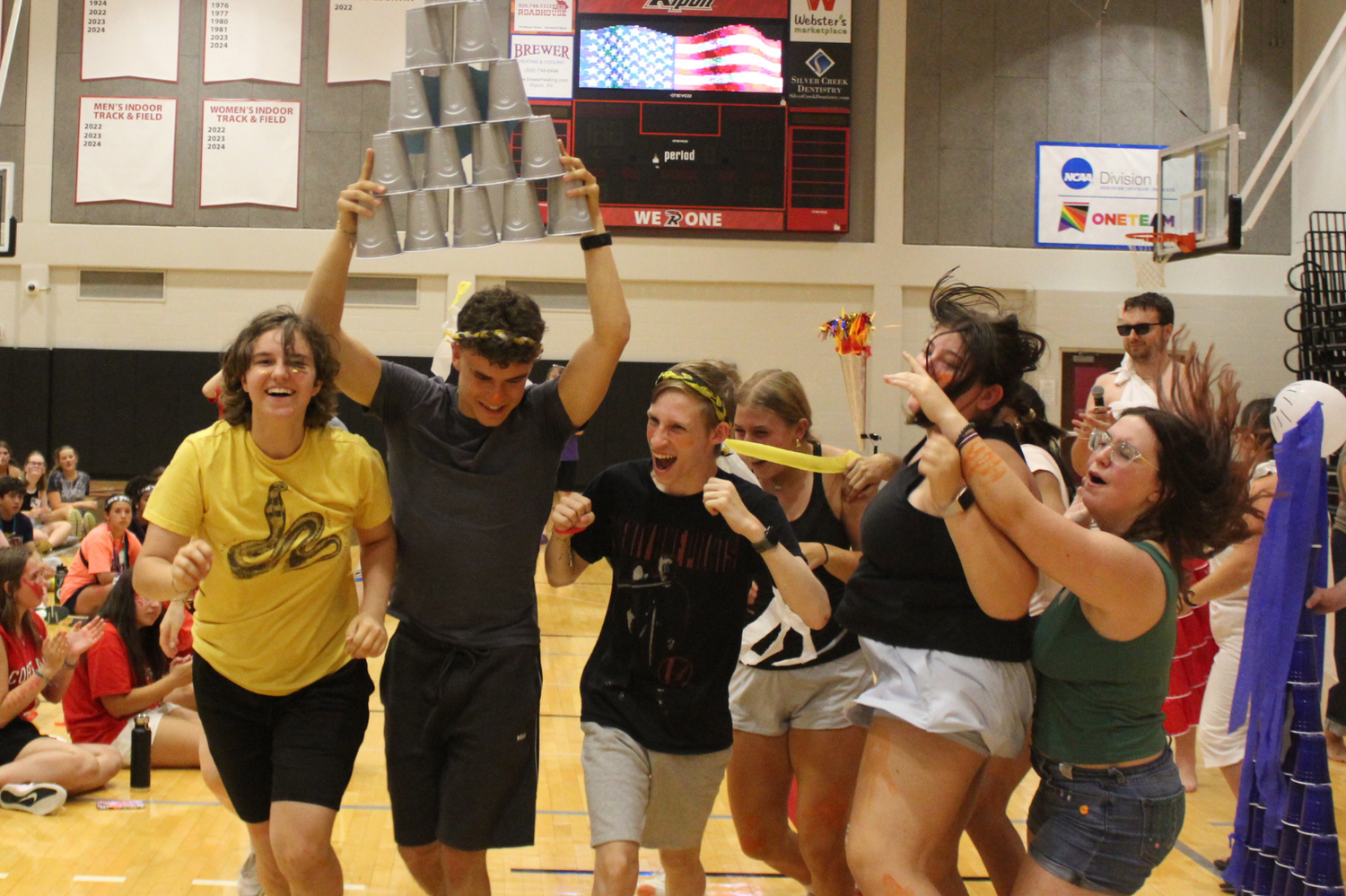 Excited group of students celebrate with raised arms during an indoor gymnasium team competition event.