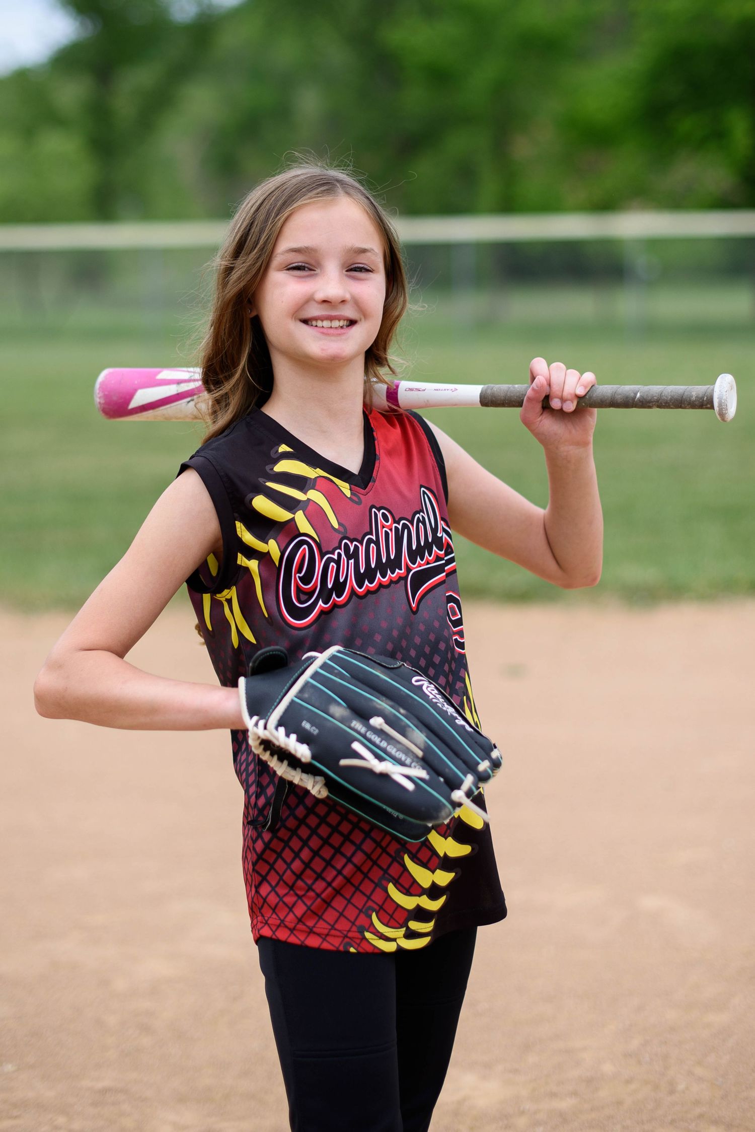 Young girl in Cardinals softball uniform smiling, holding bat over shoulder and glove, posing on baseball diamond.