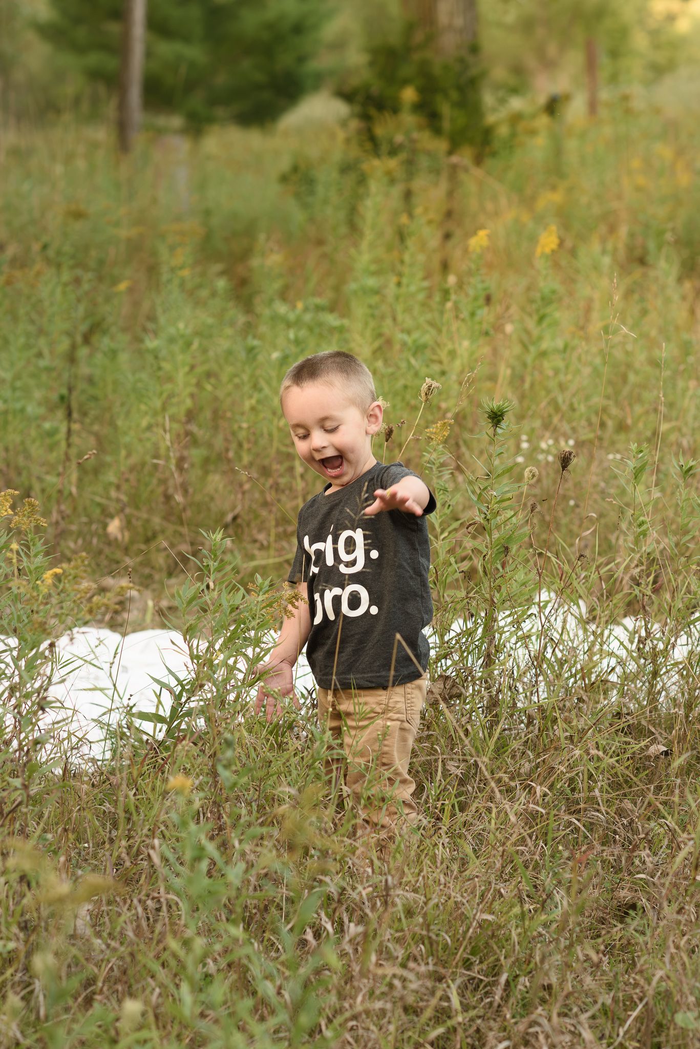 A laughing young boy in a black Big Bro shirt stands in a lush green meadow, pointing and smiling joyfully.