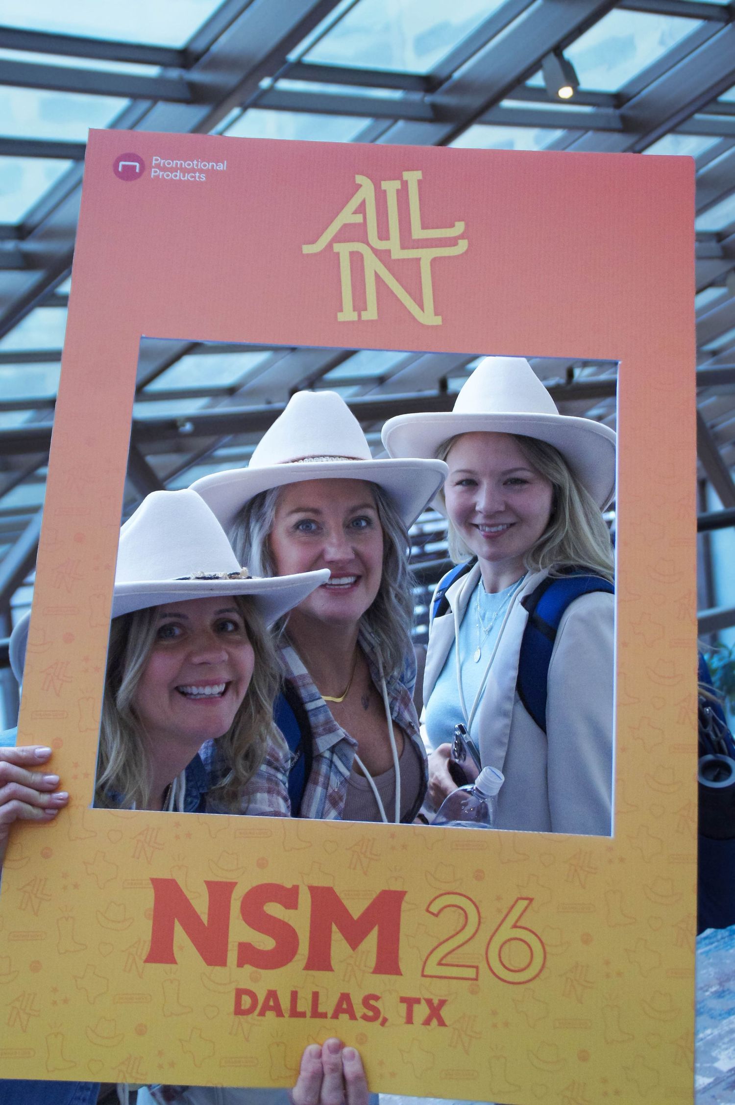 Three women in white cowboy hats pose through an All In NSM 26 Dallas TX photo frame.