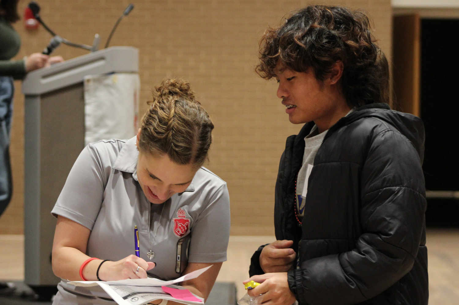 Two students collaborate at a registration table, one writing notes during a school event.