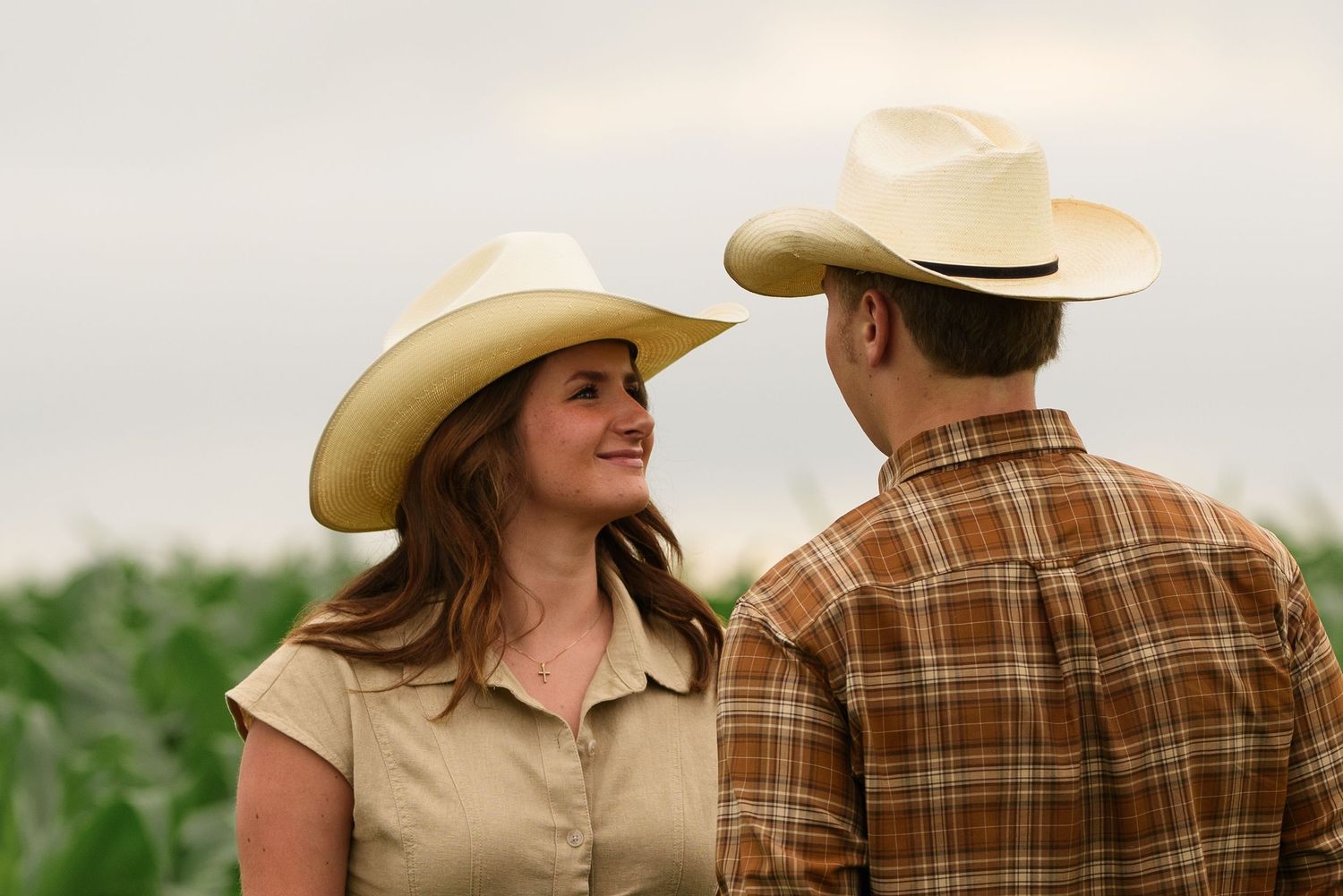 A man and woman wearing cowboy hats share a smile in a farm field with green crops in the background.