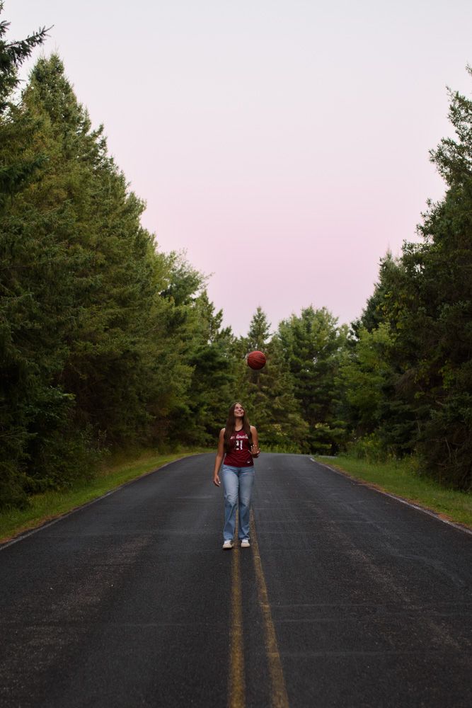 Teen girl in red jersey tossing basketball on a tree-lined road at dusk.